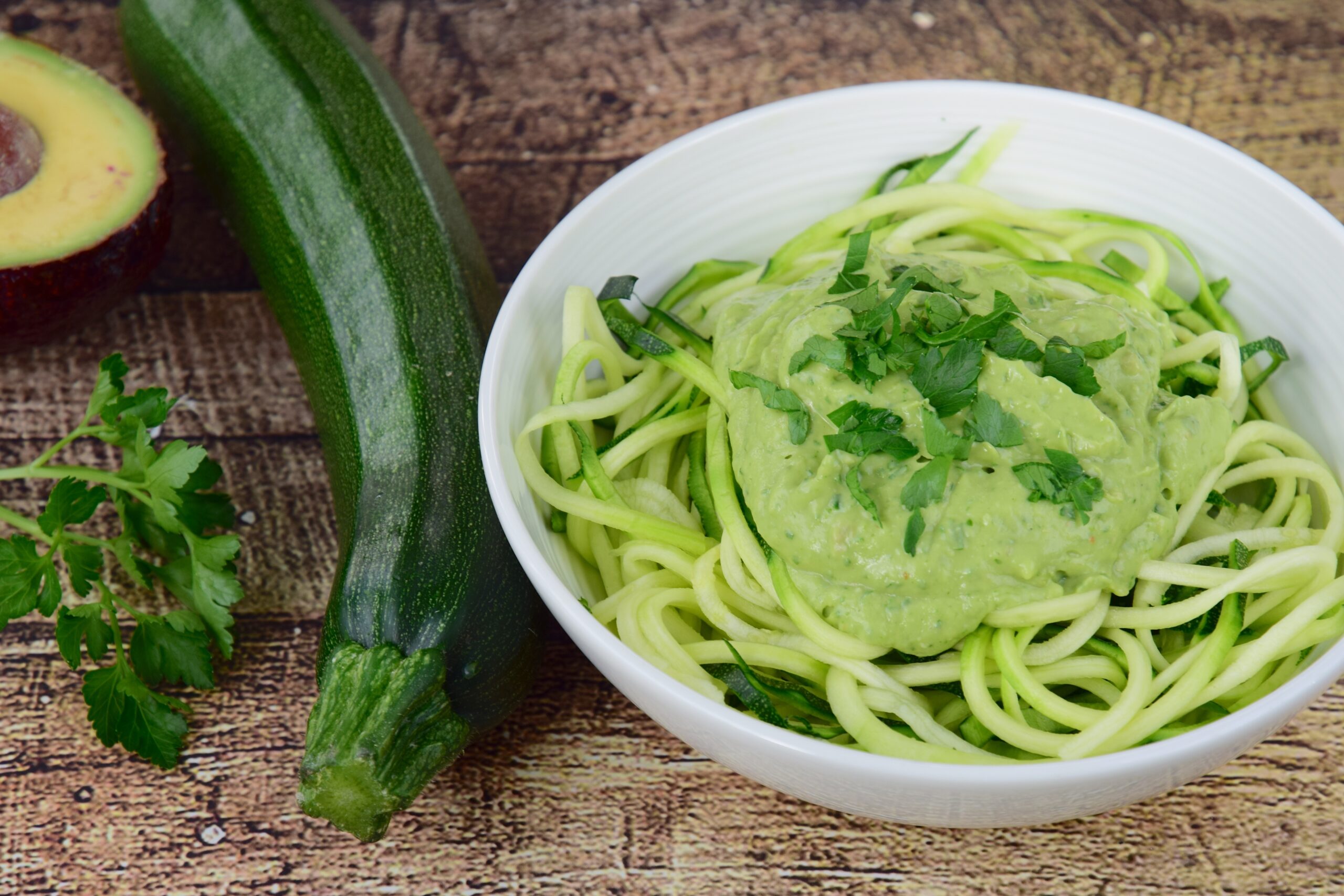 A white bowl filled with zucchini noodles topped with a creamy green sauce and garnished with fresh herbs. Next to the bowl are a whole zucchini, half an avocado, and a sprig of parsley on a rustic wooden surface.