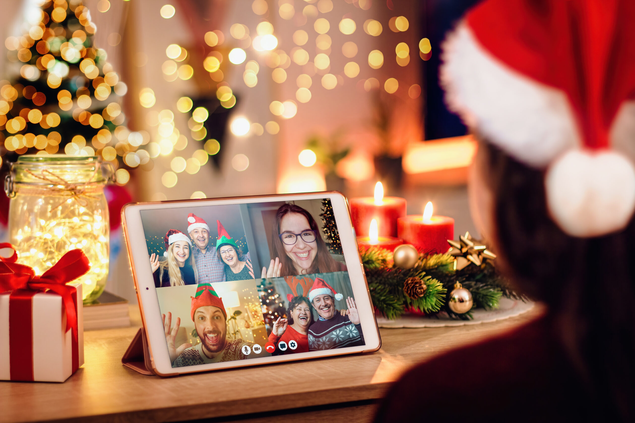 Person in a Santa hat using a tablet for a video call with four friends, all wearing festive hats. The background is decorated with Christmas lights, ornaments, candles, and gift boxes, creating a warm holiday atmosphere.