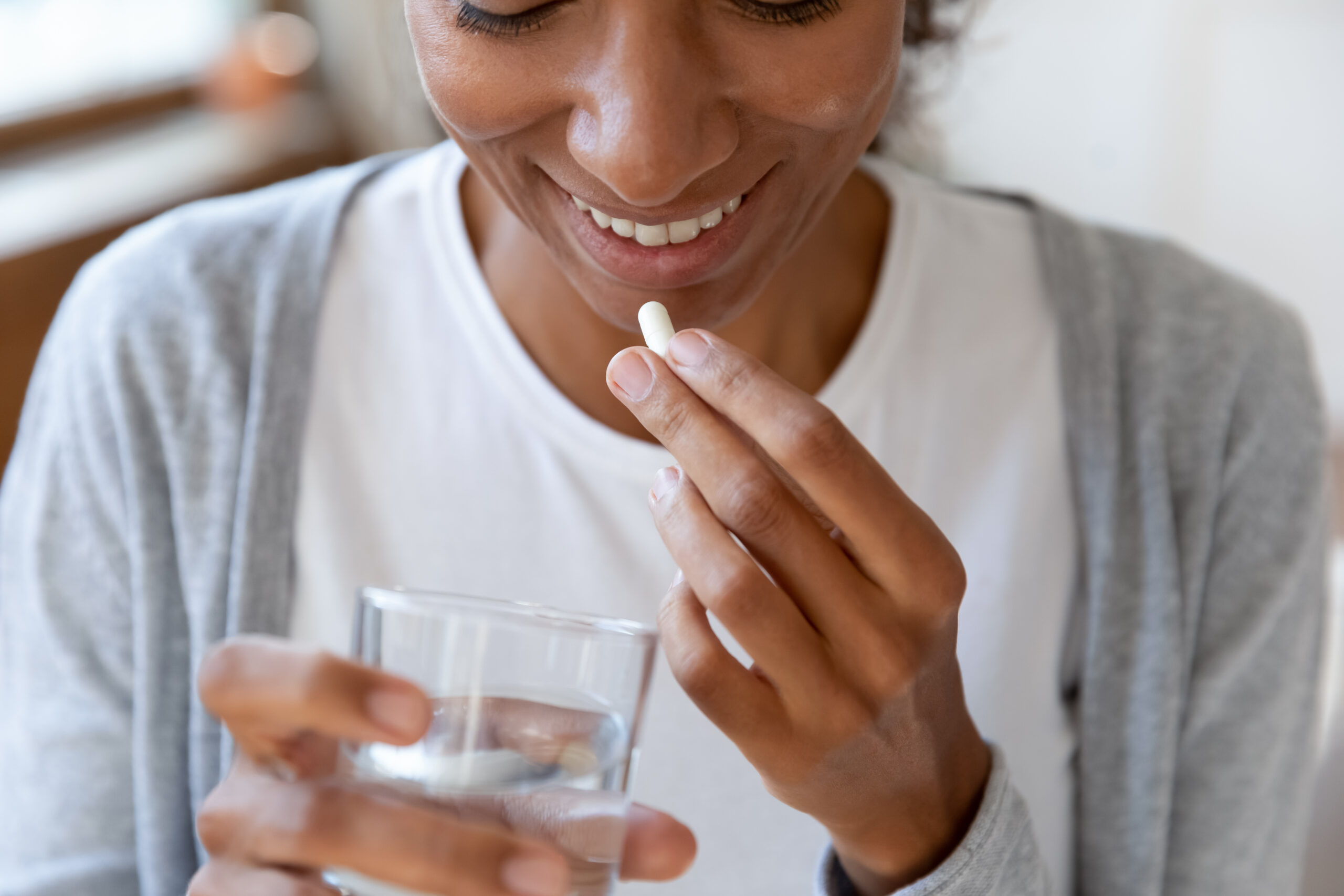 A person is holding a pill in one hand and a glass of water in the other. They are smiling, wearing a white shirt and a gray cardigan. The background is softly blurred, suggesting they are indoors, possibly near a window.