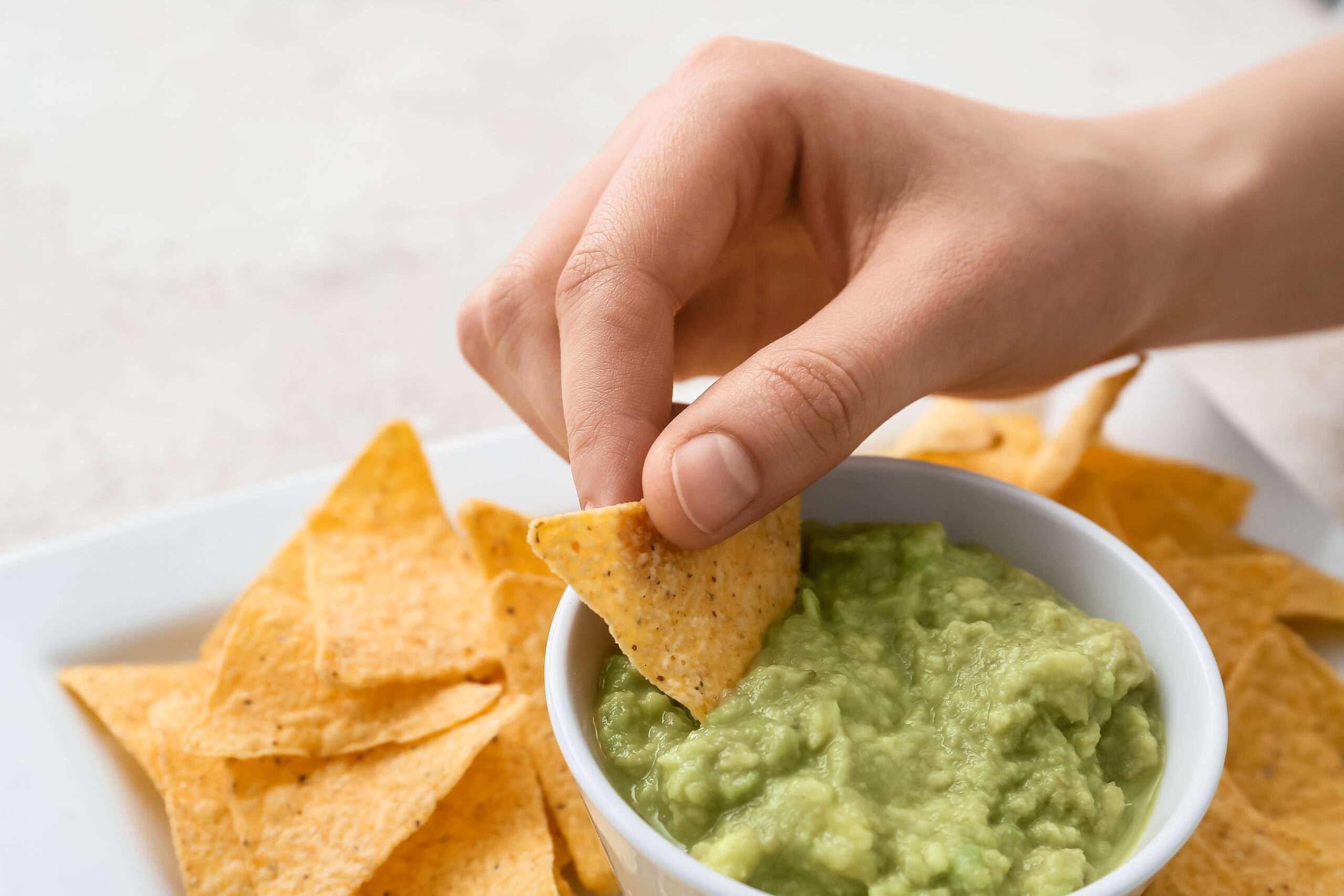 A hand is dipping a triangular tortilla chip into a bowl of creamy green guacamole. Surrounding the bowl are numerous other tortilla chips on a white surface. The background is out of focus, drawing attention to the chip and guacamole.