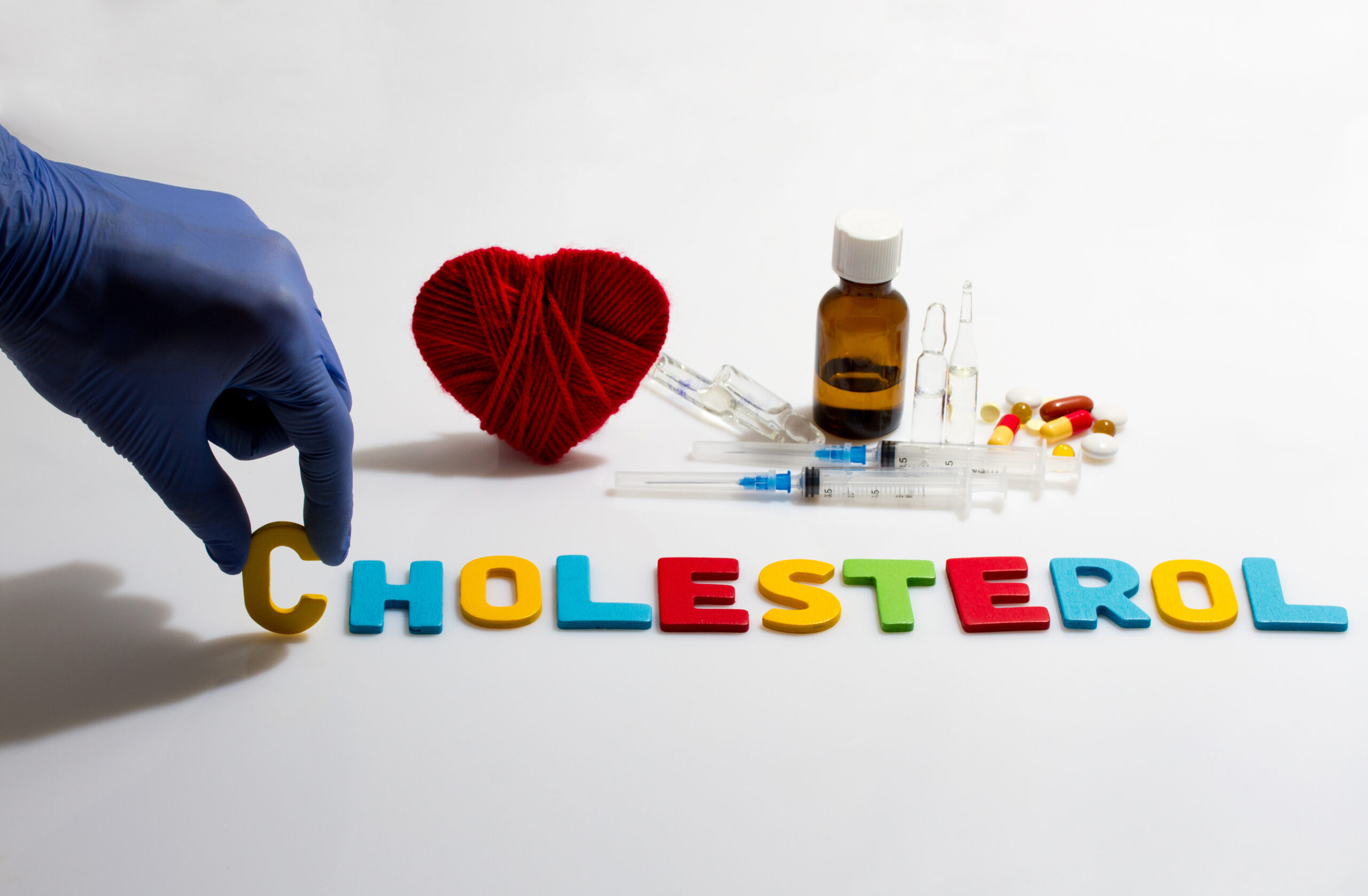 A gloved hand places a "C" next to colorful letters spelling "CHOLESTEROL." Nearby are a red yarn heart, a medicine bottle, a filled syringe, disposable syringes, and various pills on a white background.