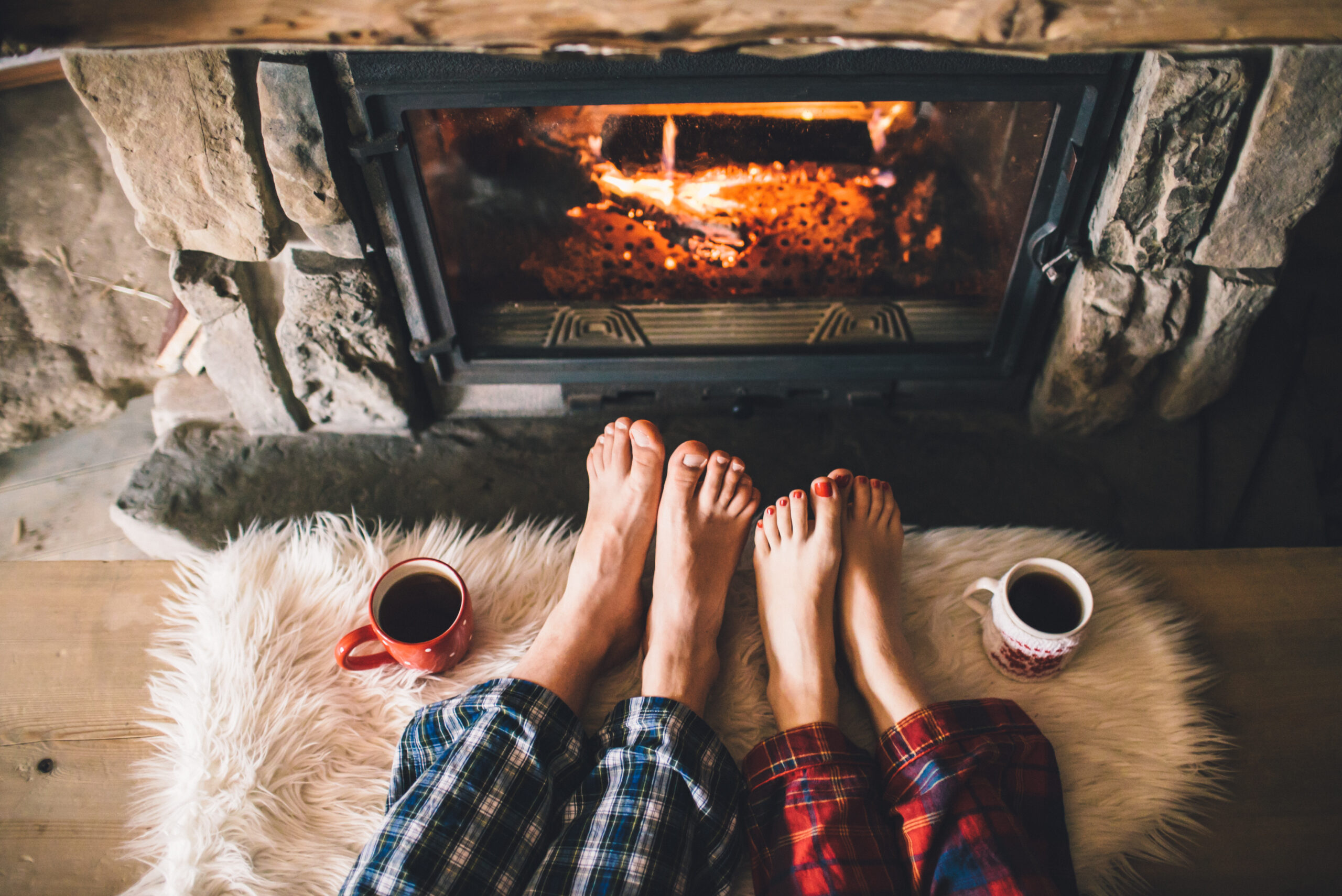 Two people are sitting with their legs outstretched, showing their feet in front of a cozy fireplace. They are wearing pajama pants, and there are two mugs of hot beverages placed on the floor beside them on a fluffy rug.