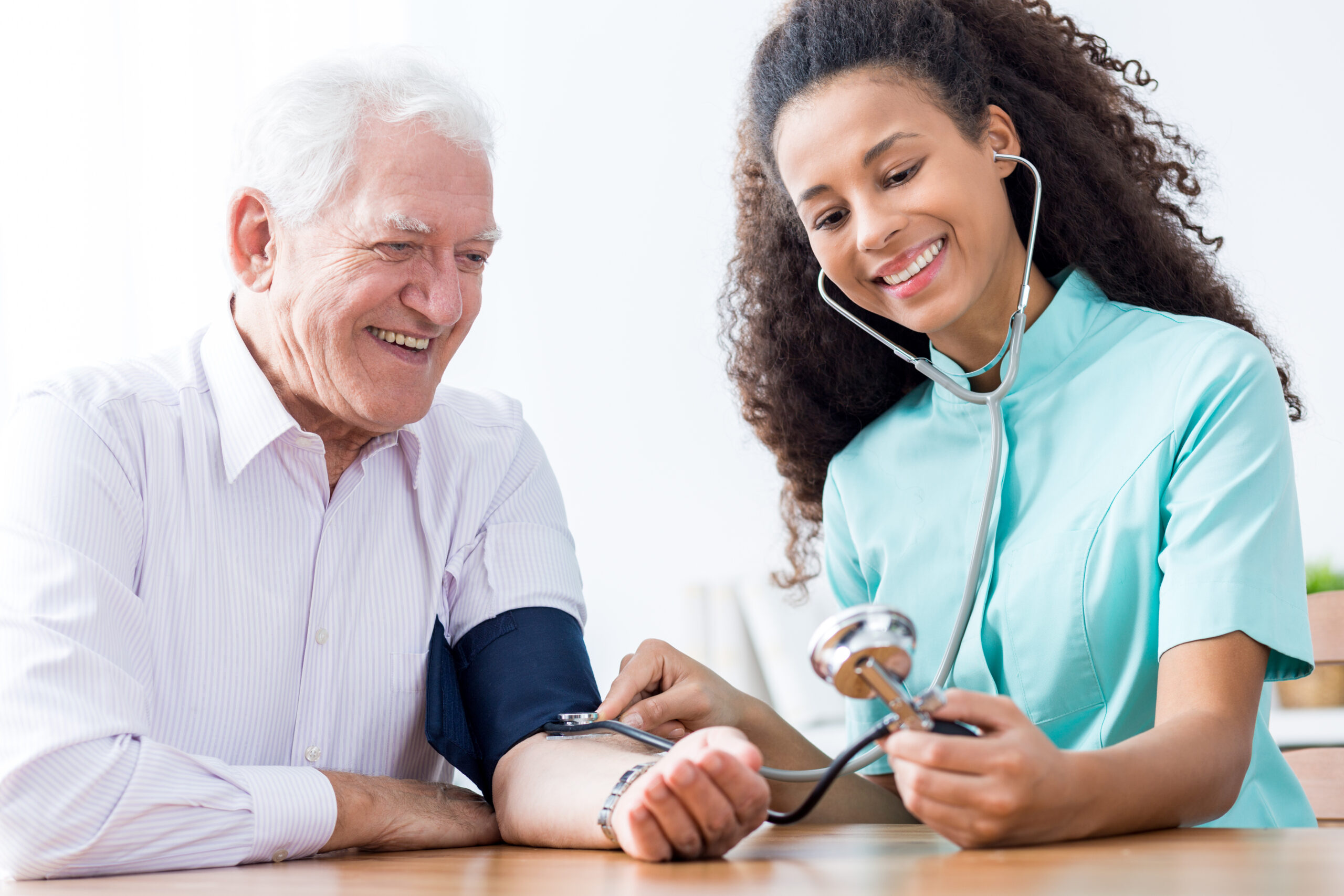 A smiling elderly man is getting his blood pressure checked by a healthcare professional. The healthcare professional, wearing a turquoise uniform, is using a stethoscope and blood pressure cuff, and she is also smiling.