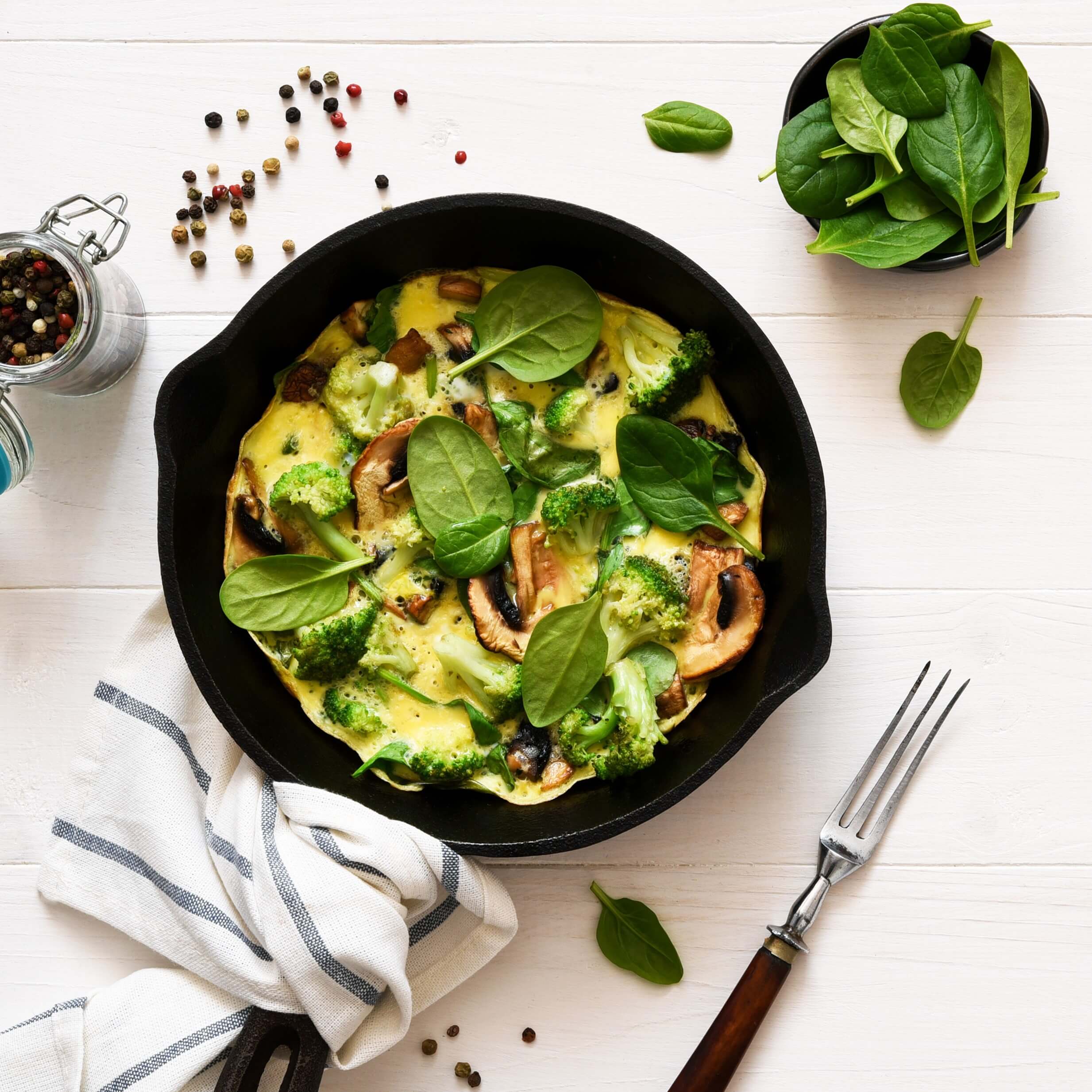 A cast-iron skillet filled with an omelette that includes mushrooms, broccoli, and fresh spinach leaves. A small bowl of spinach, a jar of mixed peppercorns, a striped kitchen towel, and a fork are arranged around the skillet on a white wooden surface.