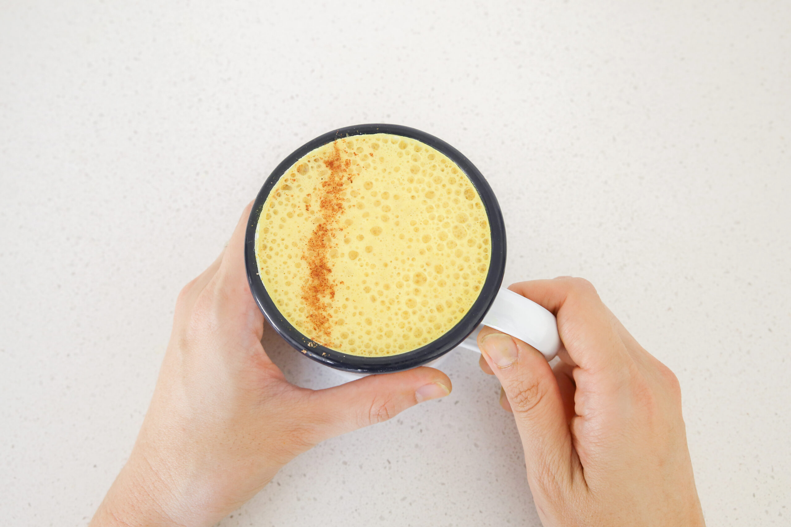 A person holding a white mug filled with a frothy yellow beverage, possibly turmeric latte, with a sprinkle of cinnamon on top. The background is a white speckled countertop.