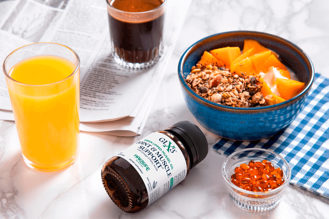 A breakfast table with a blue bowl of yogurt topped with granola and orange slices, a glass of orange juice, a glass of dark-colored liquid, a bottle of ginseng muscle support supplement, and a small dish filled with orange capsules on a blue-and-white checkered napkin.