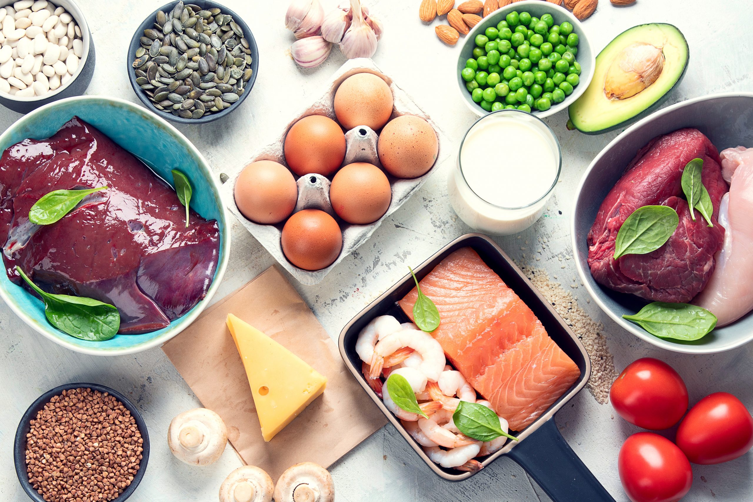 A variety of protein-rich foods are displayed on a light surface, including raw chicken breast, beef, liver, salmon, shrimp, eggs, milk, cheese, beans, peas, nuts, seeds, white mushrooms, tomatoes, garlic, and an avocado.