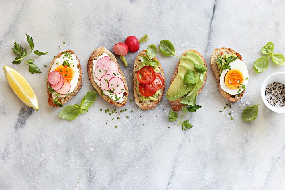 A row of five toasted bread slices topped with various ingredients: a hard-boiled egg and radish, radish slices, tomato and greens, avocado slices, and a hard-boiled egg half with herbs. Surrounding are fresh herbs, radishes, a lemon wedge, and a small bowl of salt.