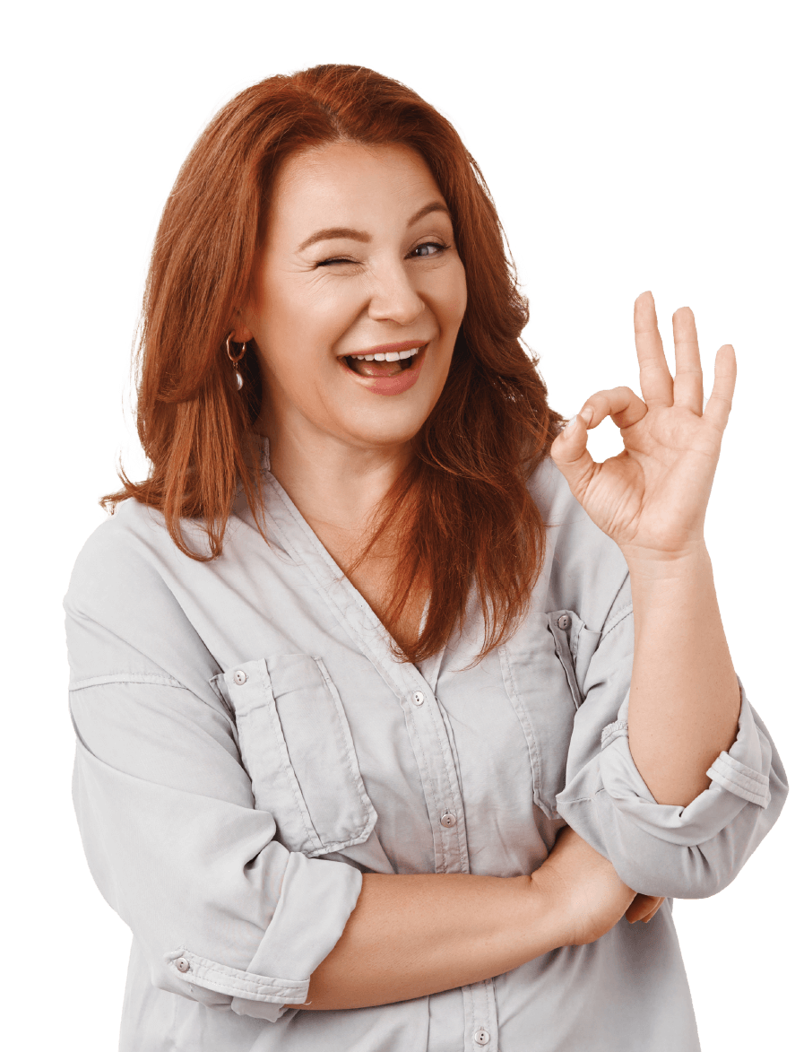 A woman with long red hair smiles and winks at the camera while giving an 'OK' gesture with her hand. She is wearing a light grey button-up shirt with pockets on the chest and has her other arm folded across her front. The background is plain white.
