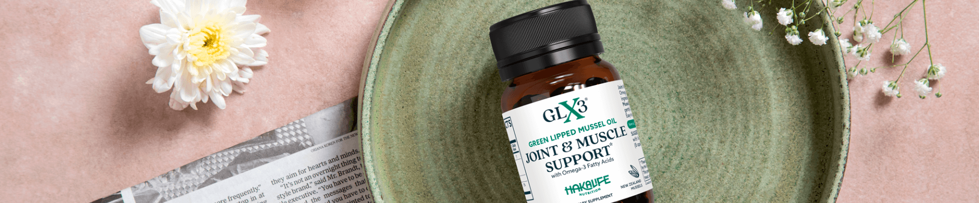 A bottle of GLX3 Joint & Muscle Support supplement, highlighting its nutritious GLX3 ingredients, is placed on a green ceramic plate. It's accompanied by a white flower and small white blossoms on a pink background, with a piece of newspaper partially visible under the plate on the left side.