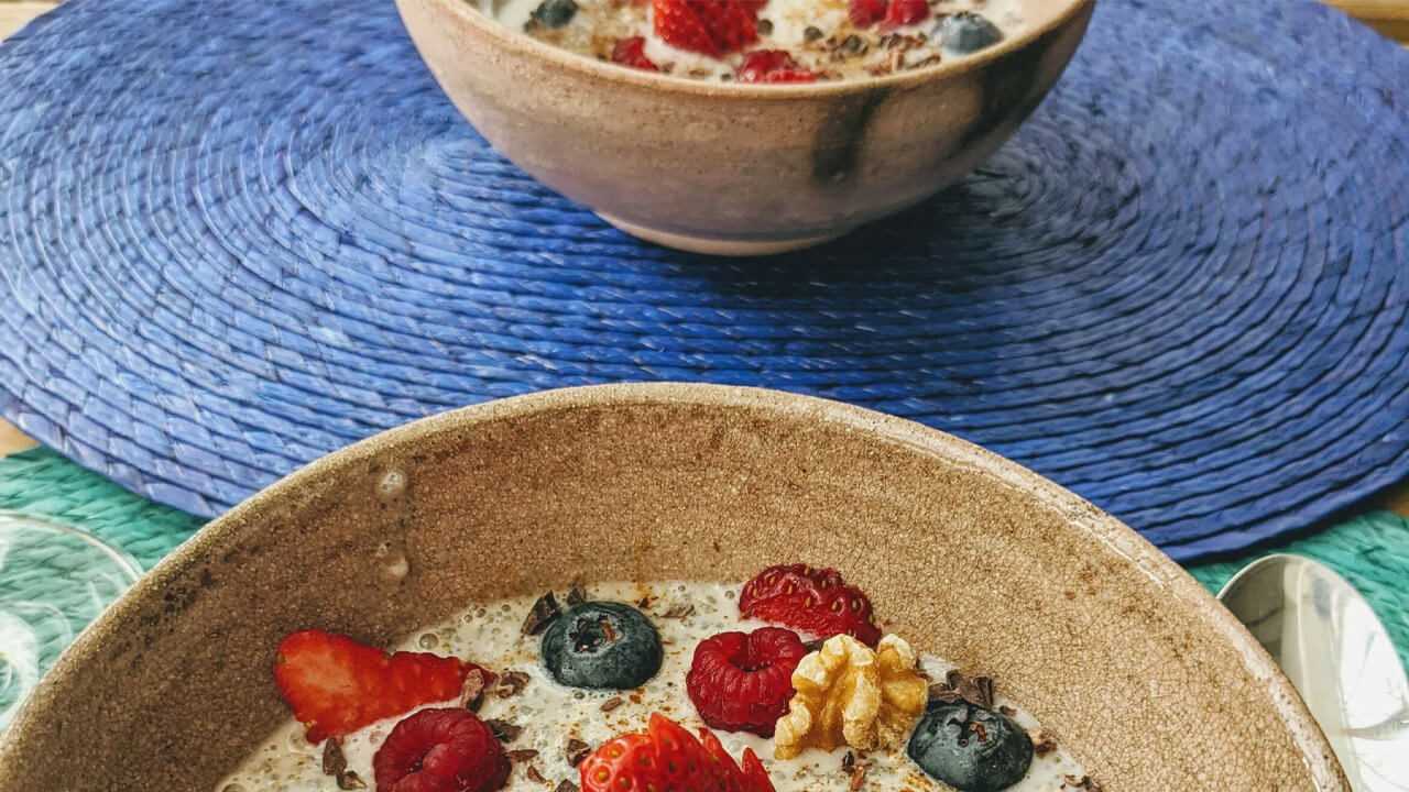 Two bowls of oatmeal topped with fresh strawberries, blueberries, raspberries, and a walnut piece are placed on a blue woven mat. The foreground shows a close-up of one bowl, while the second bowl is slightly blurred in the background.