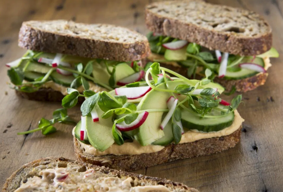 Three sandwiches made with whole grain bread, hummus, and fresh vegetables including cucumbers, radishes, and green sprouts are arranged on a rustic wooden surface. The ingredients look fresh and vibrant, showcasing a healthy, plant-based meal option.