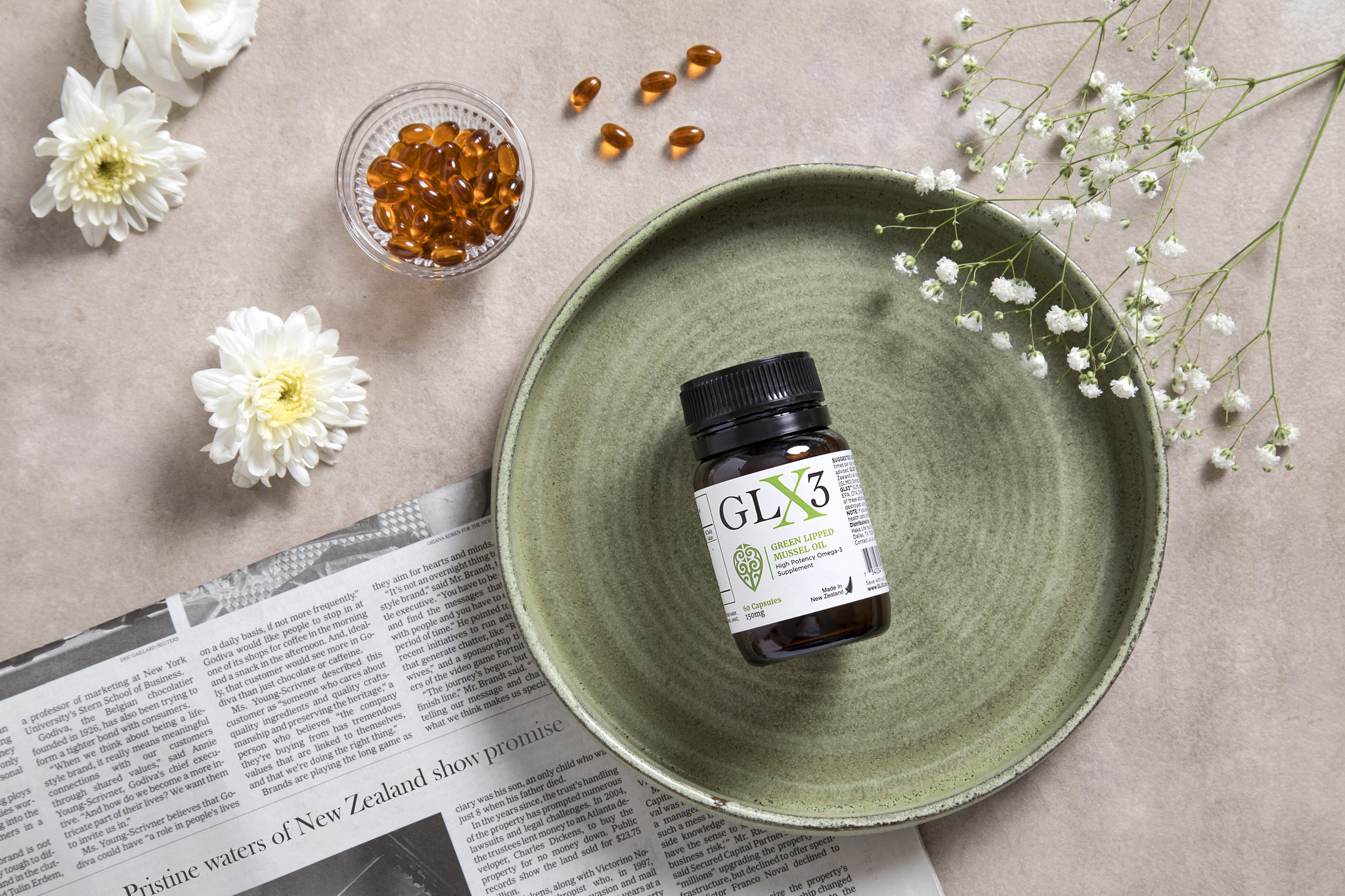 A green ceramic plate with a bottle of GLX3 supplements sits on a light-colored surface. Surrounding the plate are amber-colored capsules in a small dish, white flowers, and a newspaper. Delicate sprigs of baby's breath flowers are also visible on the plate.