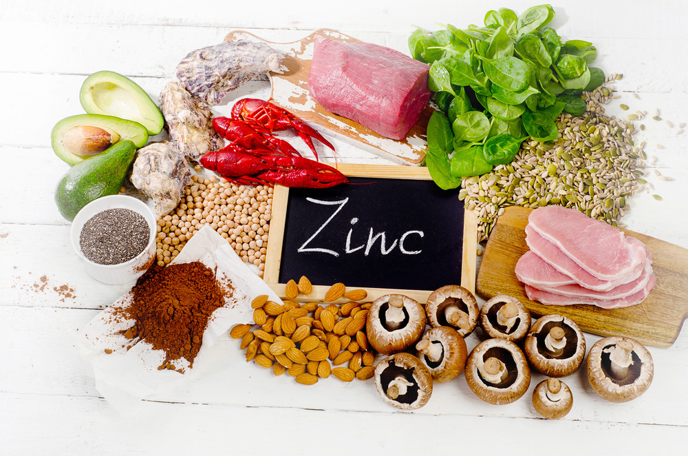 An assortment of zinc-rich foods displayed on a white wooden surface. Items include oysters, red meat, spinach, pumpkin seeds, pork, button mushrooms, almonds, cocoa powder, chickpeas, avocado, and lobster. A small chalkboard in the center reads "Zinc.