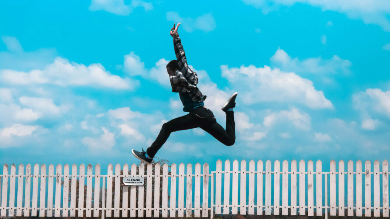A person in dark clothing is captured mid-air, jumping over a white picket fence against a backdrop of a bright blue sky with scattered clouds. This dynamic image embodies 3 things that will enable you to move freely and happy again: agility, freedom, and sheer exhilaration.