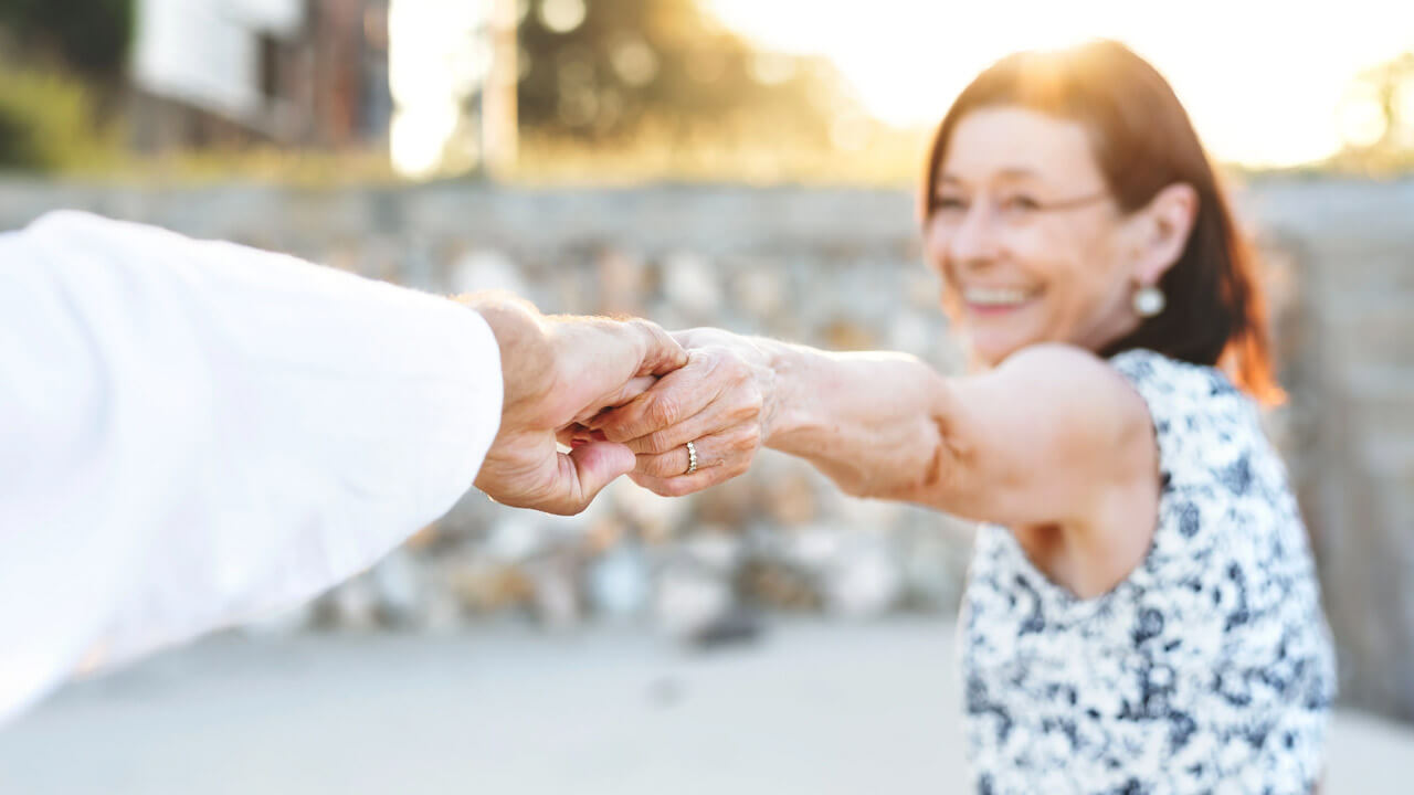A smiling woman with dark hair and a patterned dress holds hands with another person, who is partially out of frame, focusing on the hands and her joyful expression. The background is blurred with sunlight creating a warm atmosphere—truly feeling good in your fifties.