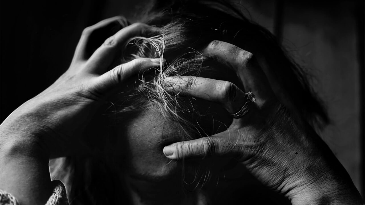 A black and white photo of a person with long hair, grasping their head with both hands in a distressed manner. The fingers are tangled in the hair, showing tension. Only part of the face and hands are clearly visible, evoking the struggles one might face before seeking simple and natural fibromyalgia treatments.