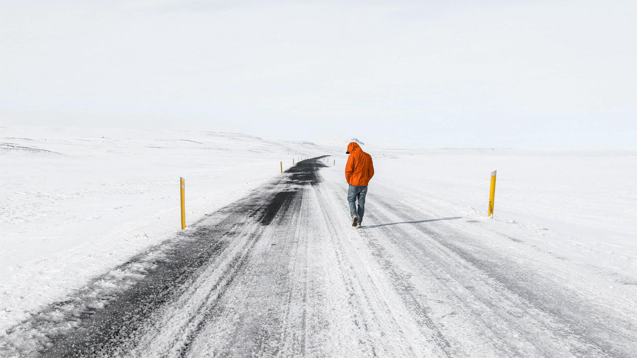 A person in an orange jacket and jeans walks down a snowy, deserted road with yellow markers on either side. The landscape is covered in snow, extending into a foggy, white horizon—an almost poetic scene that highlights the harsh weather conditions that can exacerbate arthritis pain.