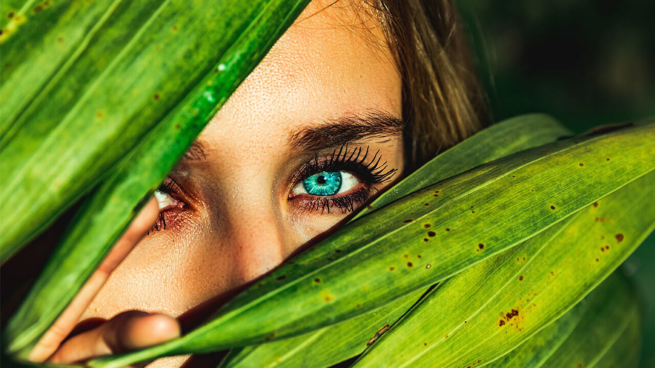 A close-up of a woman's face partially obscured by large green leaves. One blue eye, benefiting from omega-3, is visible, looking directly at the camera and framed by dark eyelashes. The vibrant green leaves dominate the foreground, adding a natural element to the image.