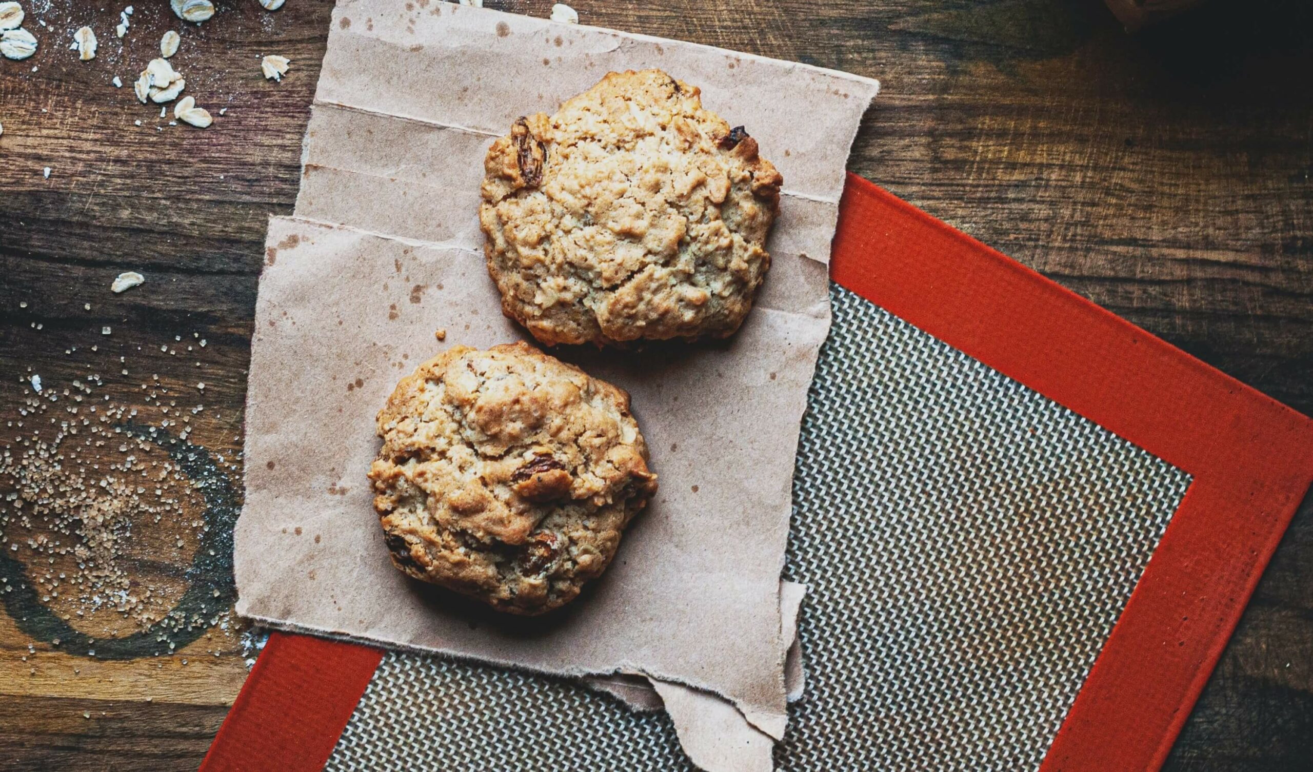 Two oatmeal cookies rest on a piece of brown parchment paper. They are placed on a wooden surface with a red and beige silicone baking mat partially visible in the background. Some scattered oats and crumbs are also visible around the cookies.