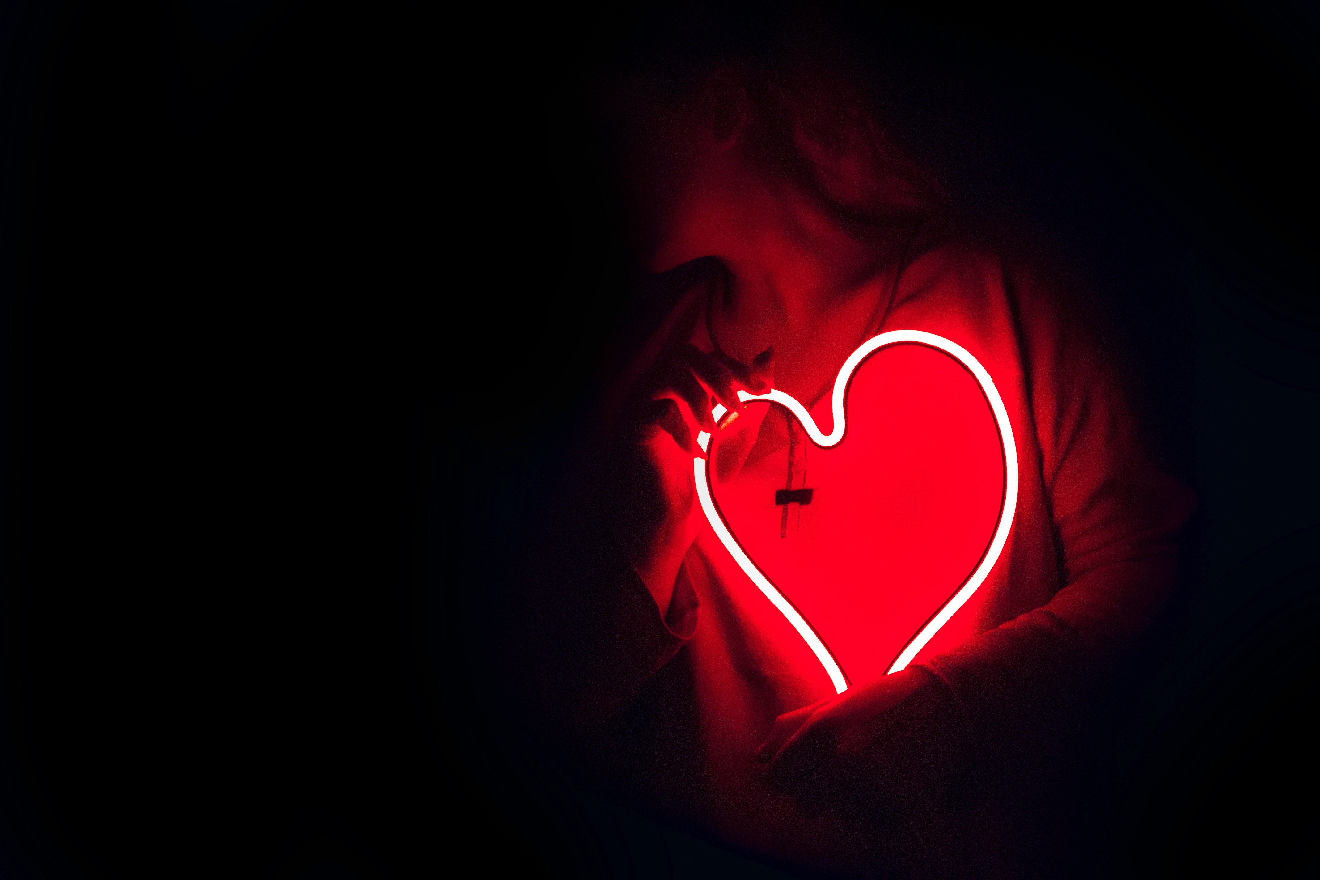 A person in low light holds a bright red neon heart, akin to asking, "Can omega-3 reduce heart attacks?" The glowing heart illuminates part of their face and upper body, casting a soft red light. The background is almost completely dark, focusing attention on the person and the heart.