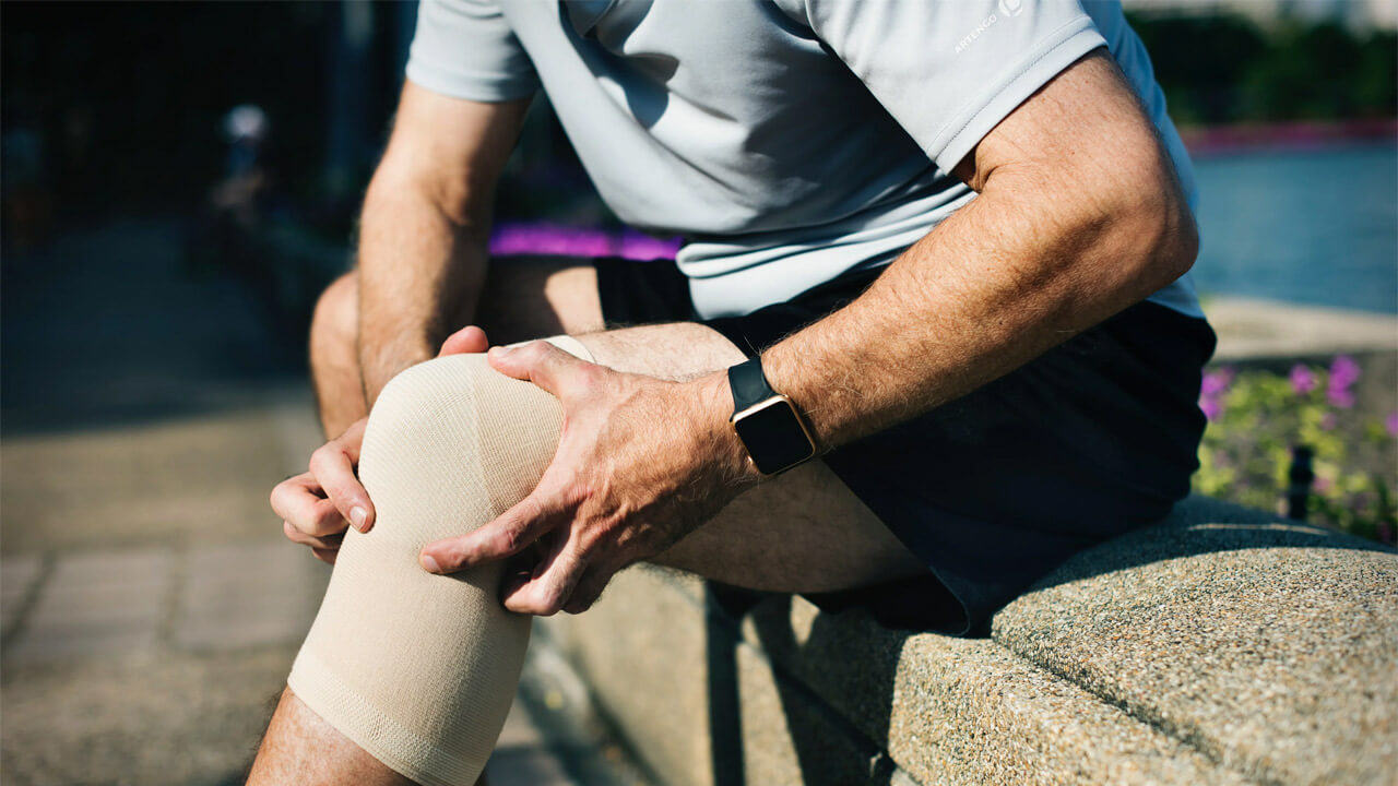 A person wearing a light blue shirt and black shorts is sitting on a stone bench outdoors, their knee wrapped in a beige compression bandage. They are wearing a smartwatch on their left wrist, possibly pondering the connection between arthritis and sports amidst the blurred urban background.