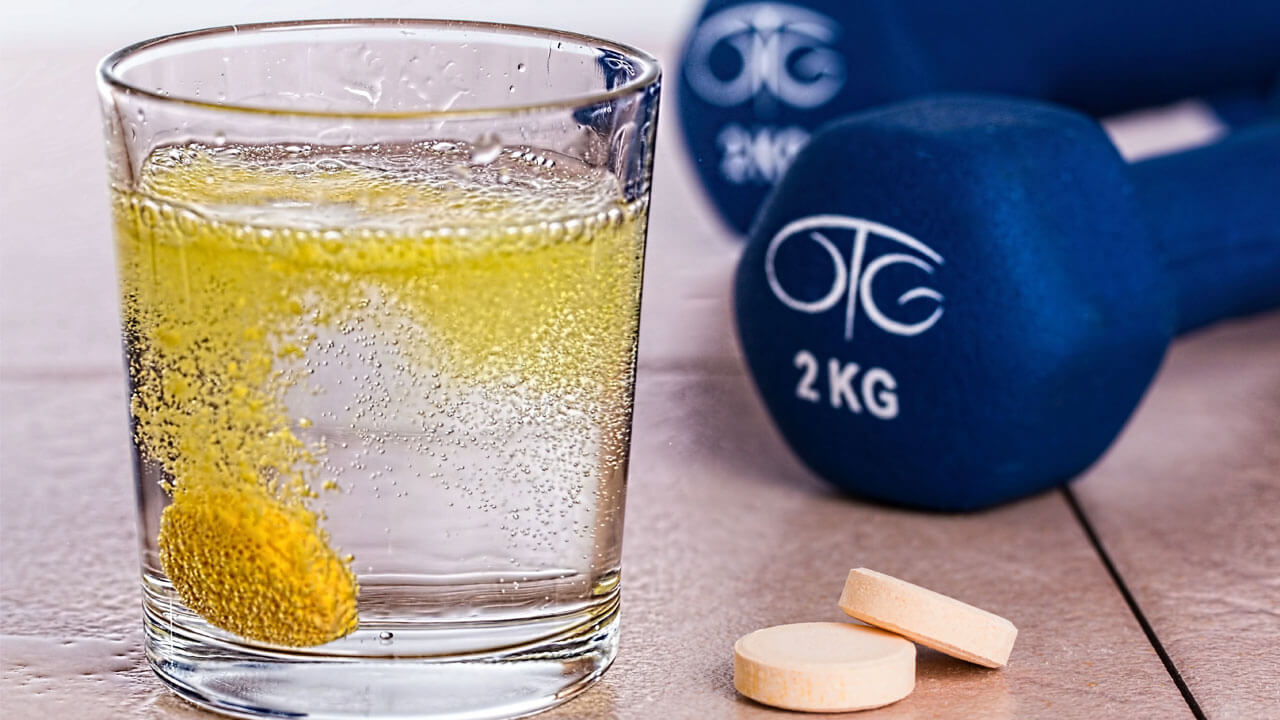 A dissolving yellow effervescent tablet fizzes in a glass of water next to two more tablets on a table. In the background, there are two blue 2-kilogram dumbbells embossed with white logos, emphasizing the importance of vitamins for joint pain relief.