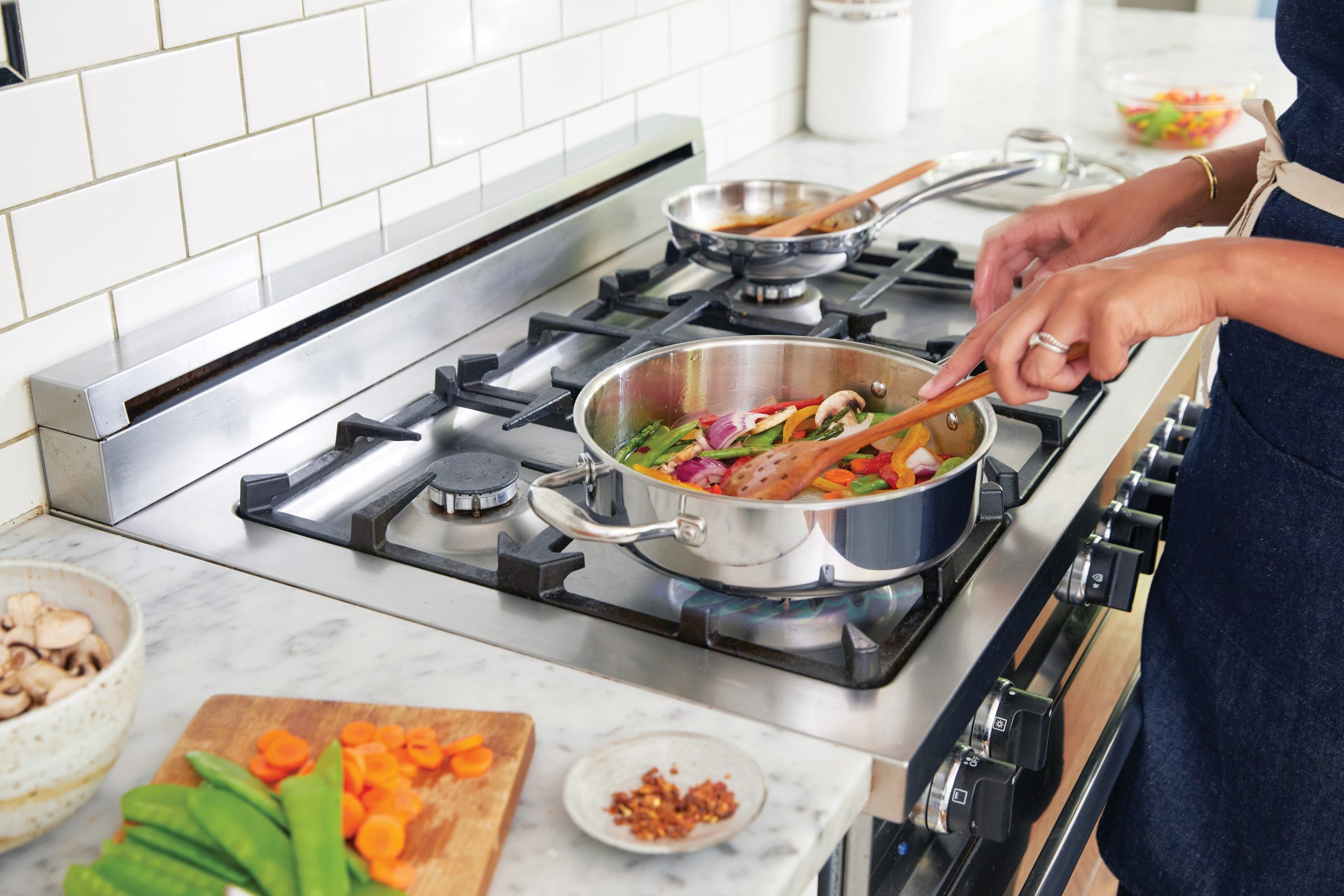 A person wearing an apron cooks vegetables in a stainless steel pan on a gas stove. Ingredients such as sliced carrots, snow peas, and mushrooms are on the marble countertop nearby. The background features a white subway tile backsplash.