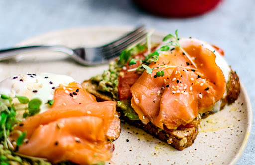A close-up of a dish featuring two pieces of toast topped with sliced smoked salmon, avocado spread, poached eggs, and sprinkled with black and white sesame seeds. A fork is visible in the background on a lightly speckled plate.