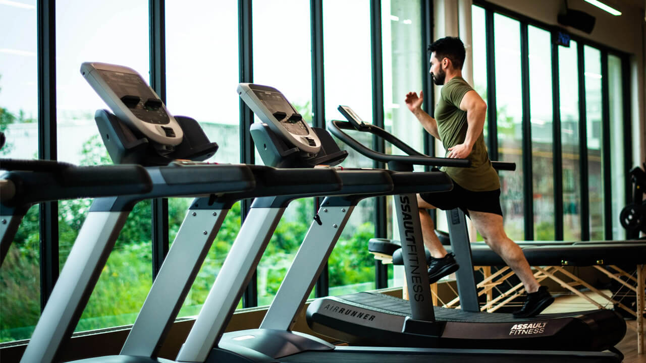 A man with a beard is running on a treadmill in a gym with large windows, embracing natural ways to strengthen bones and joints. The gym has multiple treadmills, and the man is focused on his workout, wearing a green shirt and black shorts. Greenery outside can be seen through the windows.