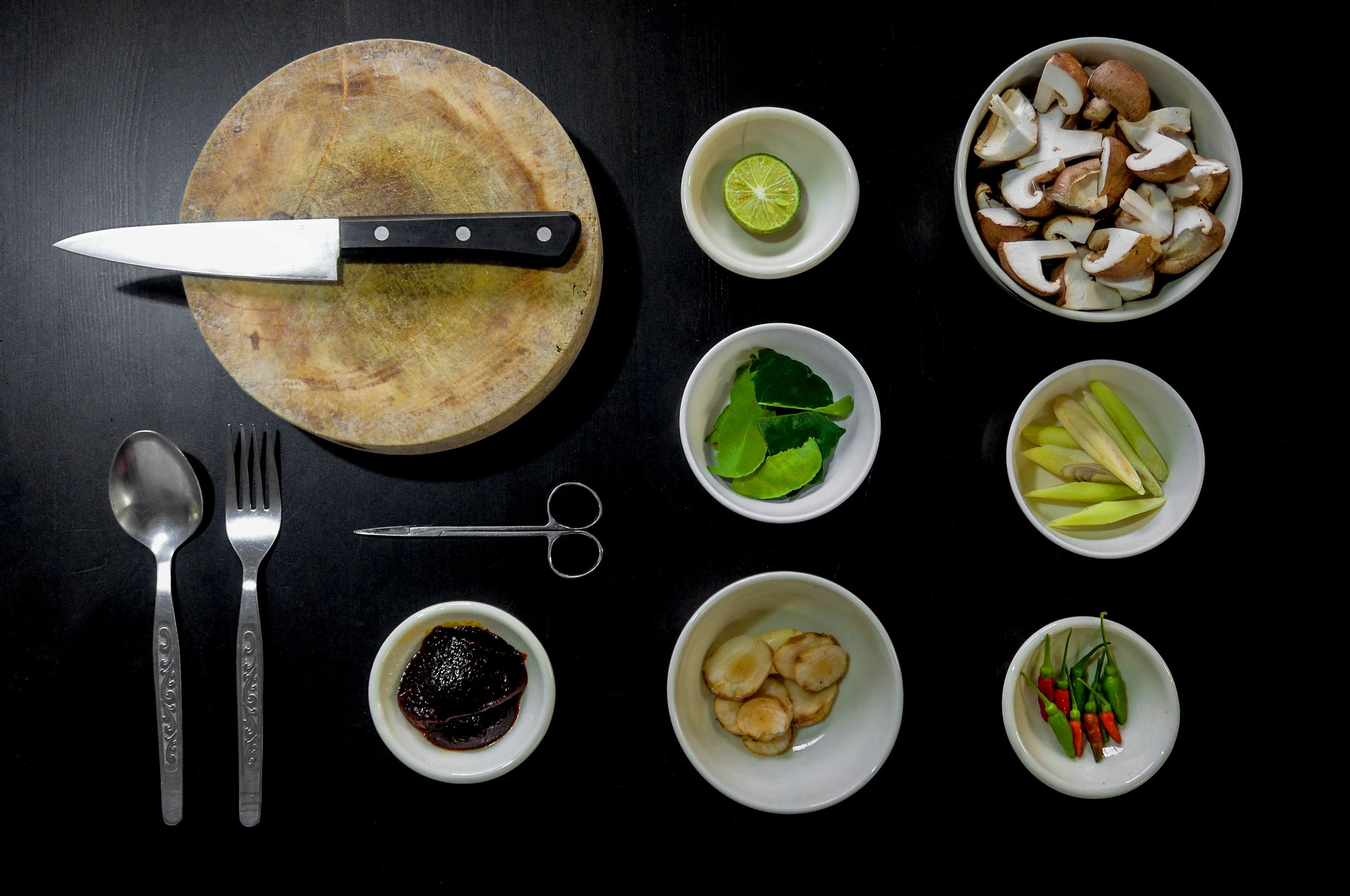 A top-down view of cooking ingredients and utensils arranged neatly on a dark surface. Items include a knife on a wooden cutting board, a spoon, a fork, kitchen scissors, a lime, mushrooms, kaffir lime leaves, lemongrass, garlic, chili peppers, and black sauce—perfect for easy meals to make with arthritis.