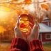 Hands in a red sweater hold a jar with a maple leaf inside, against a background of vibrant autumn leaves and a quaint red and white house. The warm sunlight filters through the foliage, creating a cozy, nostalgic atmosphere.