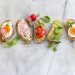 A row of five toasted bread slices topped with various ingredients: a hard-boiled egg and radish, radish slices, tomato and greens, avocado slices, and a hard-boiled egg half with herbs. Surrounding are fresh herbs, radishes, a lemon wedge, and a small bowl of salt.