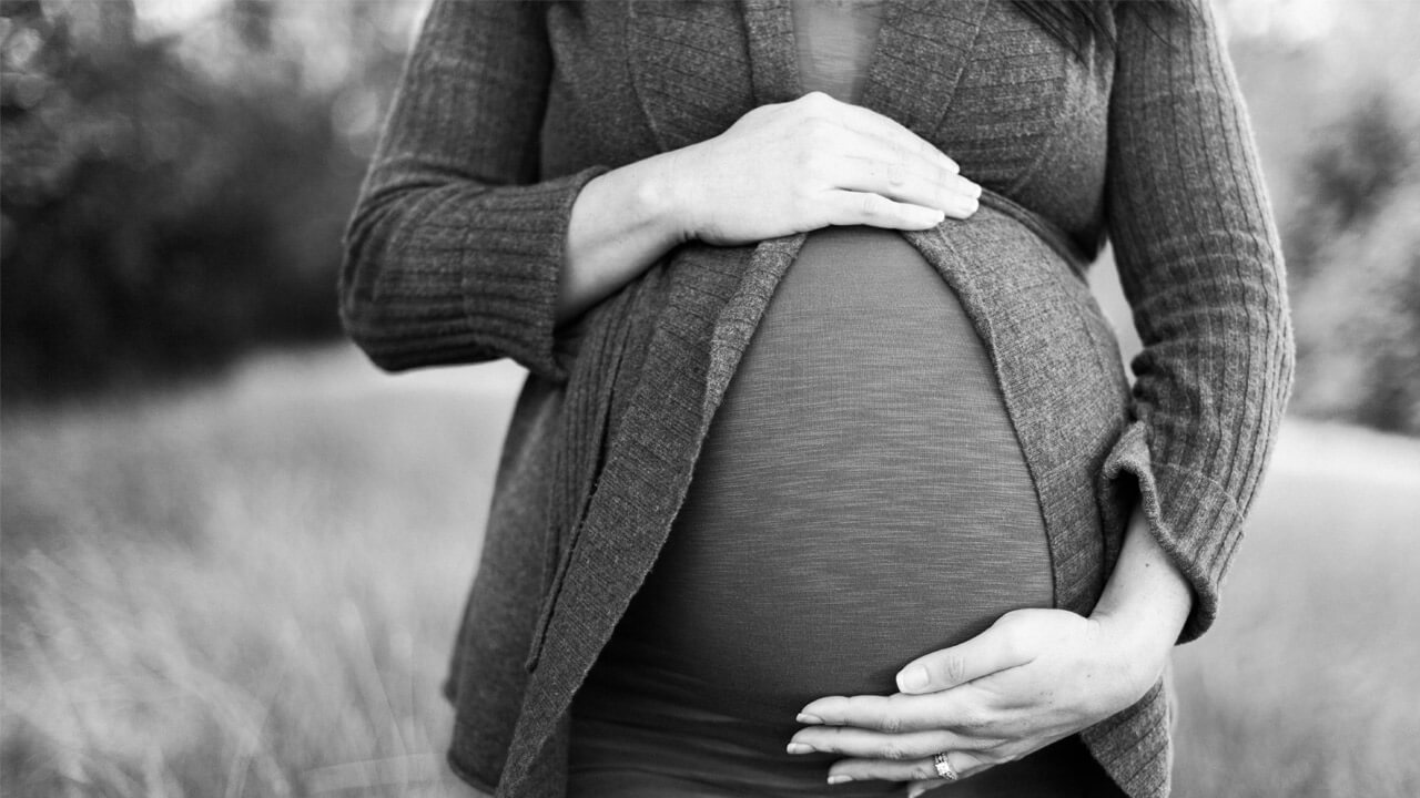 A black-and-white image showing a pregnant woman cradling her large belly with both hands. She is wearing a cardigan over a fitted top, and her face is not visible. The background, blurred with natural outdoor scenery, evokes thoughts of safe foods for pregnancy and maternal care.