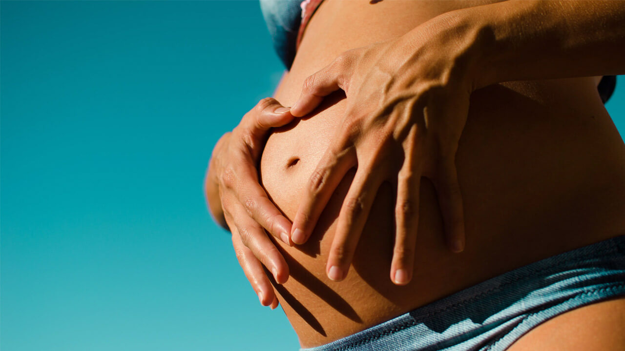 Close-up of a person’s bare pregnant belly, cradled by their hands. The skin is sunlit, and the background is a clear blue sky. The person's fingers are interlocked over the belly, creating a heart shape—a reminder of why Omega-3s might be good for your pregnancy.
