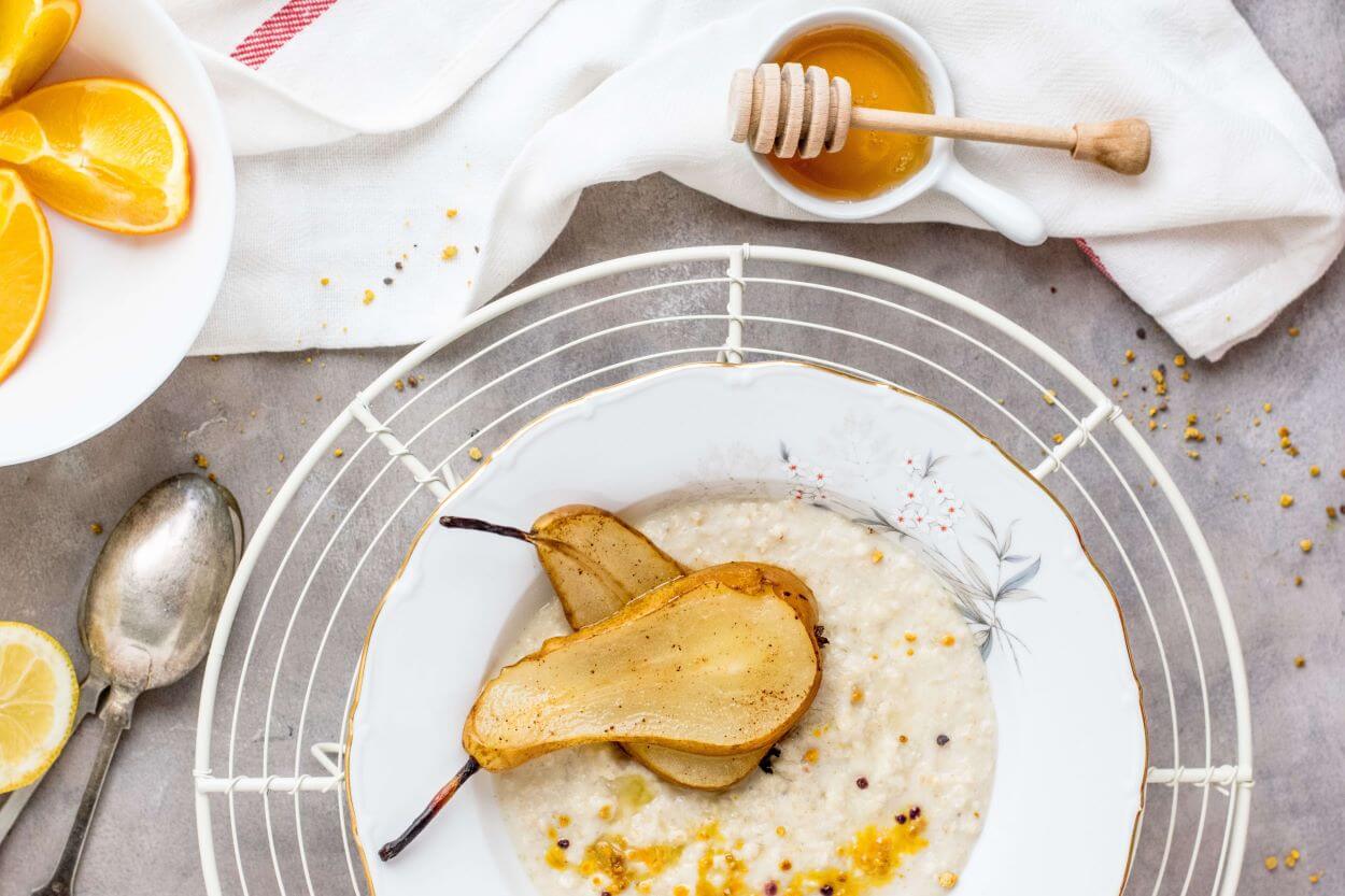 A plate of porridge topped with roasted pear halves sits on a white tablecloth. Next to the plate are a small bowl of honey with a honey dipper, and a plate of orange slices. A vintage spoon and a lemon wedge are also visible in the scene.