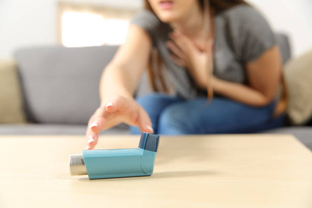 A person reaching for a blue asthma inhaler on a table while holding their chest, clearly in distress. The background is slightly blurry, showing a couch and part of a window, highlighting the urgency as they reach for relief.