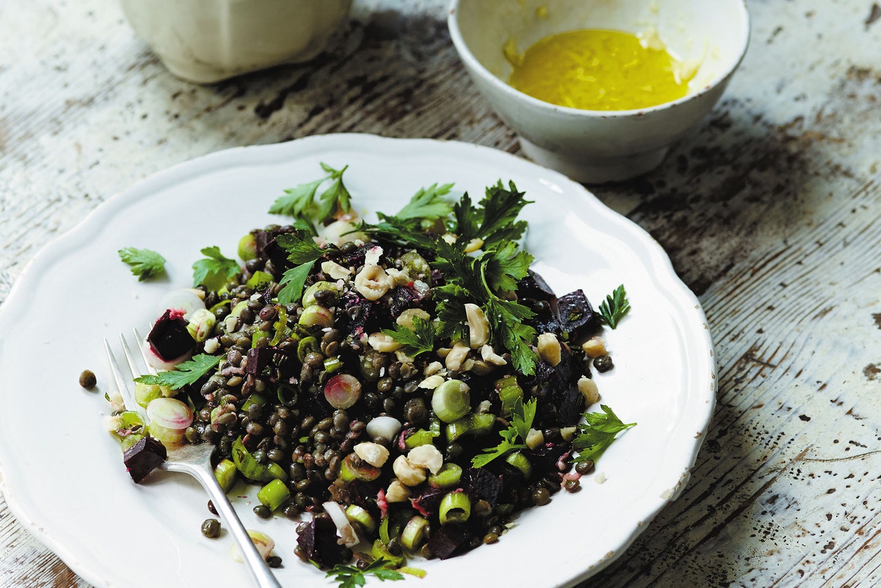 A plate of mixed lentil salad with chopped vegetables, herbs, and nuts is displayed on a rustic wooden table. Next to it is a bowl of yellow dressing. A fork is placed on the plate, ready for use.
