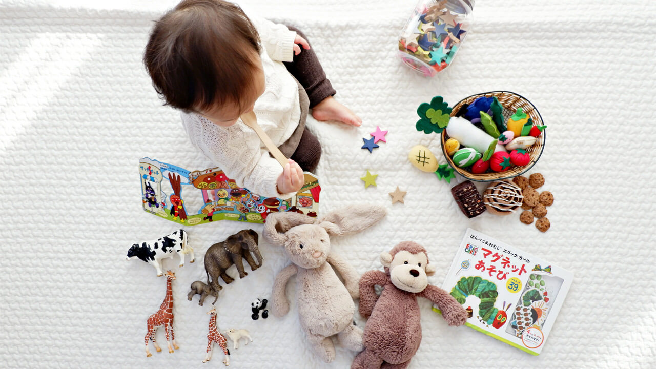 A baby sits on a white blanket surrounded by various toys, including stuffed animals, plastic animals, colorful wooden stars, and picture books. The baby is reading a colorful book with animal illustrations. Nearby, alongside a jar of craft supplies, lies a pamphlet titled "Shocking New Study Suggests Omega-3 Benefits Your Kid’s Behavior.