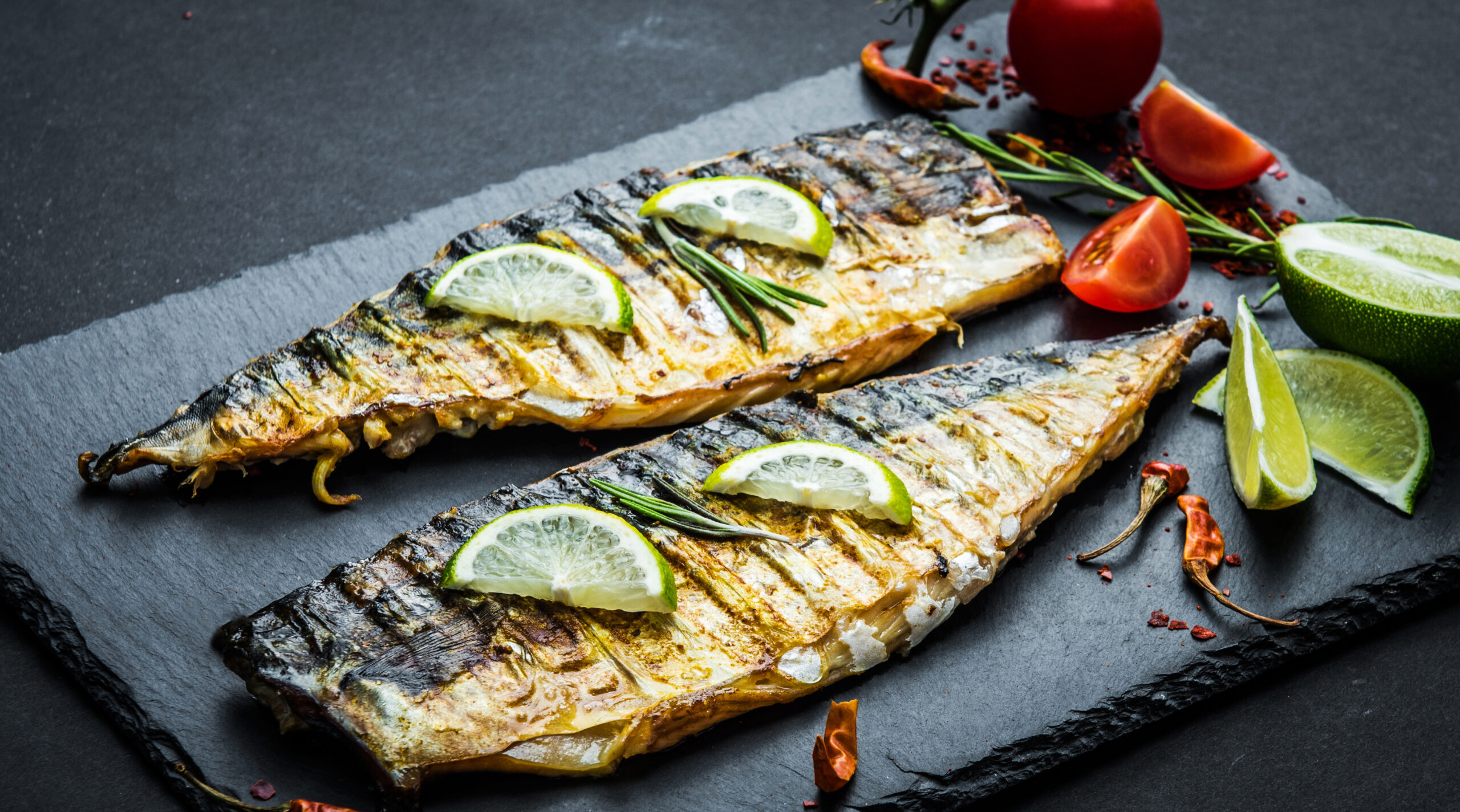 Two grilled fish fillets garnished with lime wedges and rosemary sprigs are placed on a dark slate plate. Surrounding the fish are halved limes, cherry tomatoes, and scattered chili peppers, creating a vibrant and appetizing presentation.