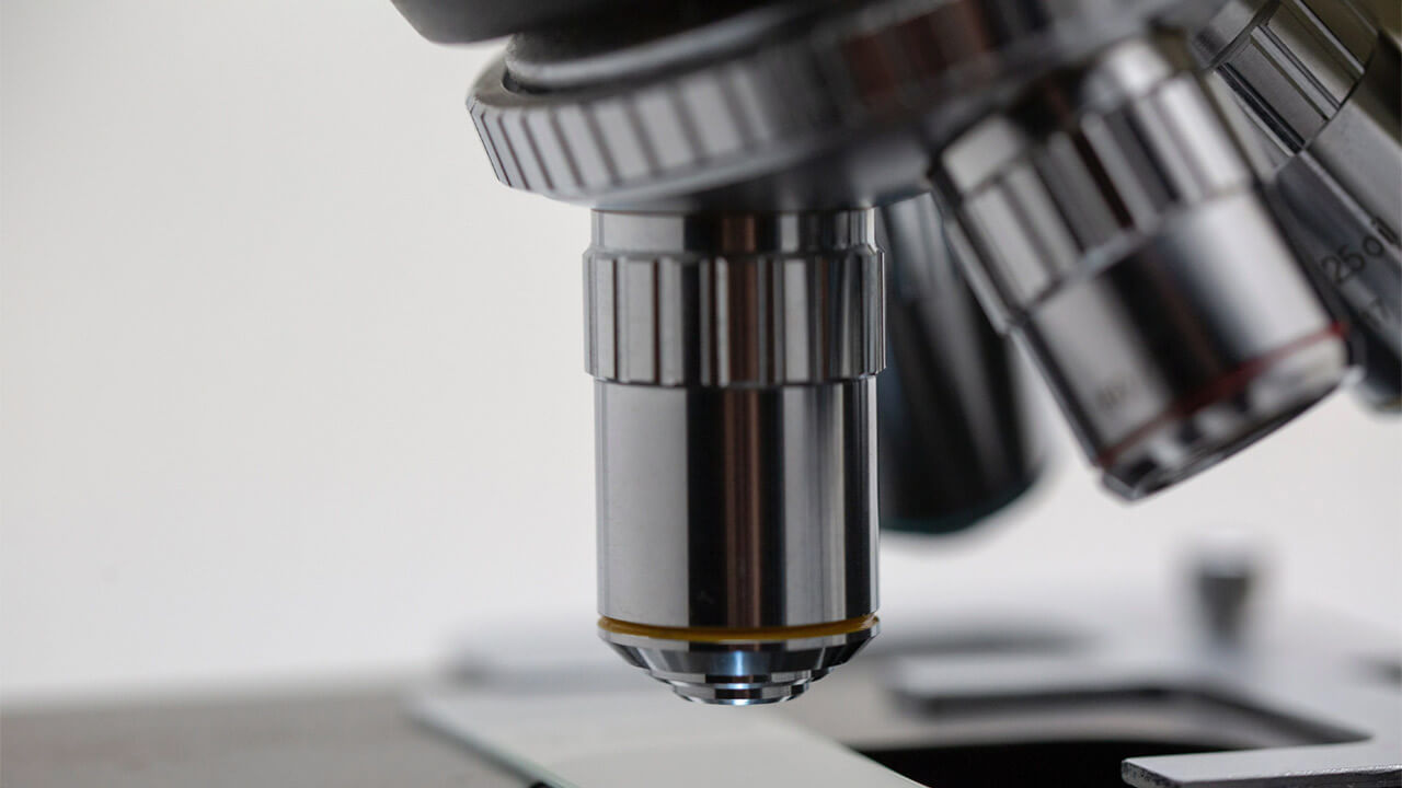Close-up image of the objective lenses of a microscope. The lens in focus has a metallic finish and ridged edges for adjustment, reminiscent of the precision seen in ETA omega 3 chain mechanisms. The background is slightly blurred, highlighting the fine details of the microscope's components.