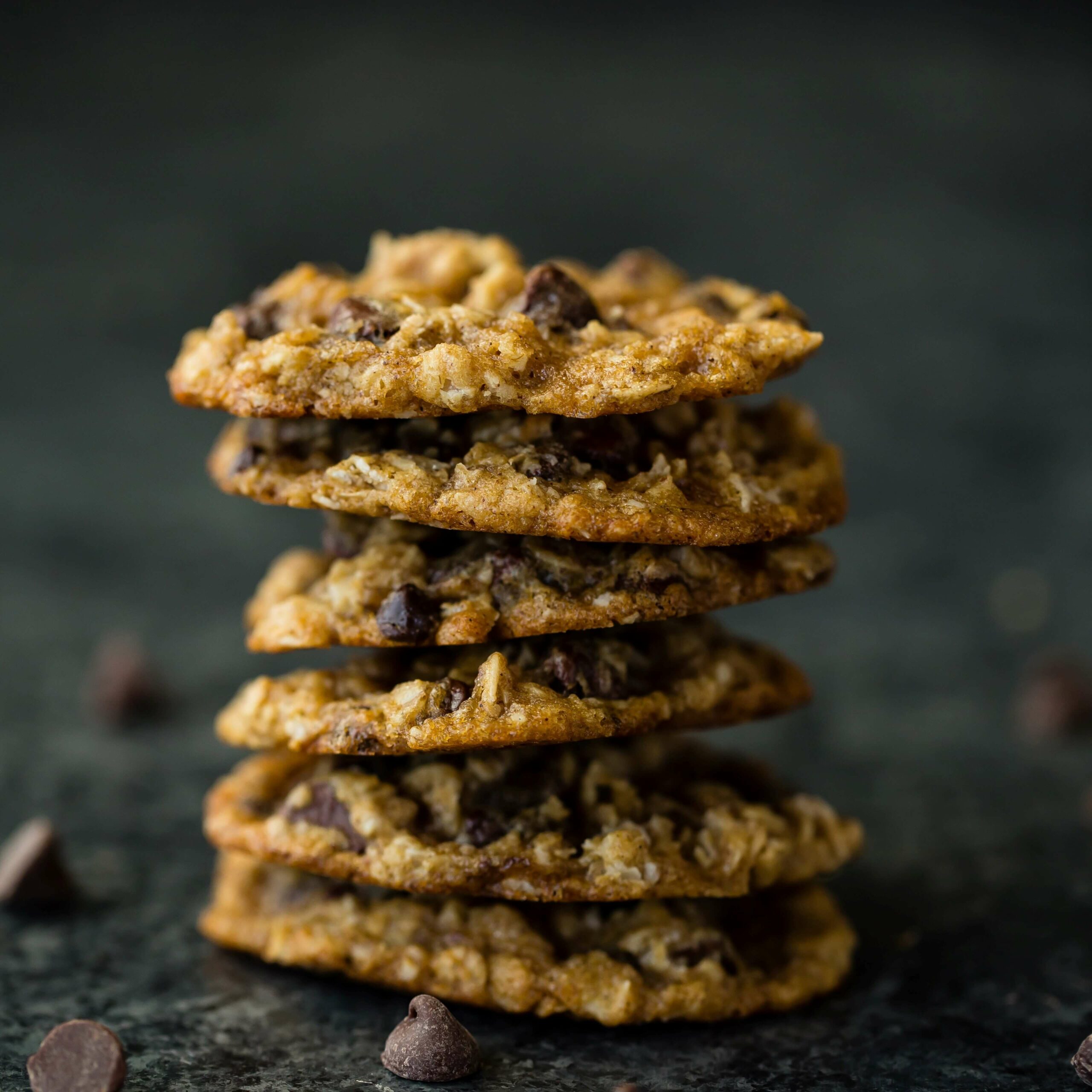 A stack of five chocolate chip oatmeal cookies is placed on a dark, textured surface. Several chocolate chips are scattered around the base of the stack, adding to the inviting, homemade feel of the scene. The cookies look crispy on the edges and chewy in the center.