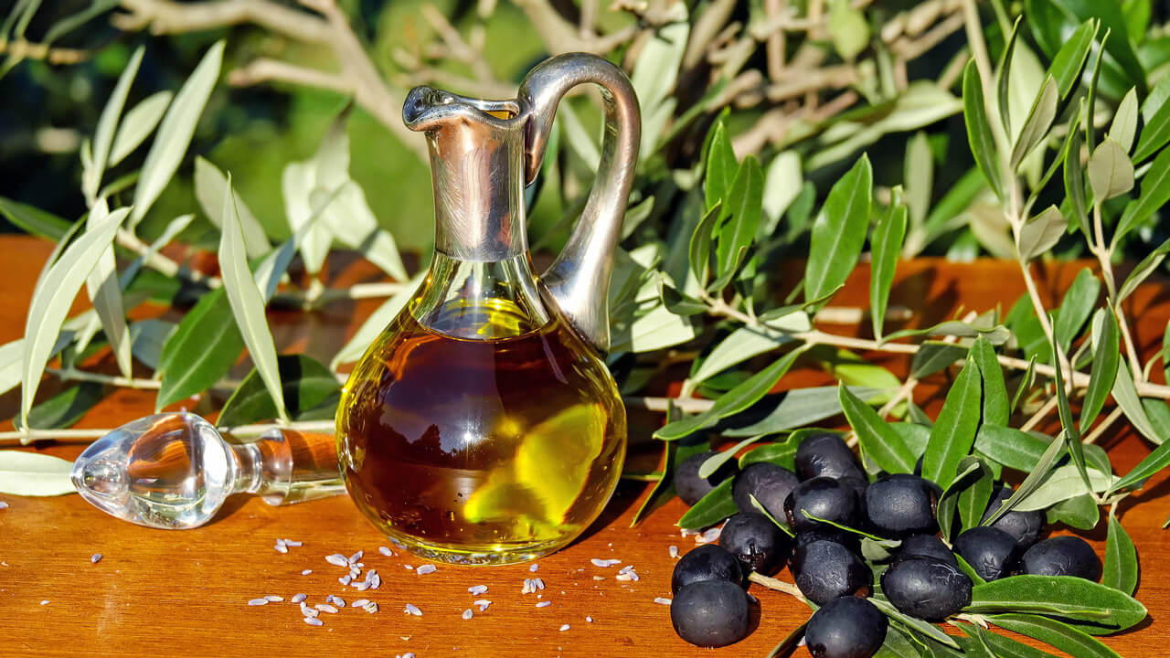 A glass jug filled with olive oil sits on a wooden surface, surrounded by fresh olive branches and a bunch of black olives. A decorative glass stopper lies nearby, and sunlight highlights the scene, showcasing the benefits of New Zealand Olive Oil and why we use it to create such a fresh and natural atmosphere.