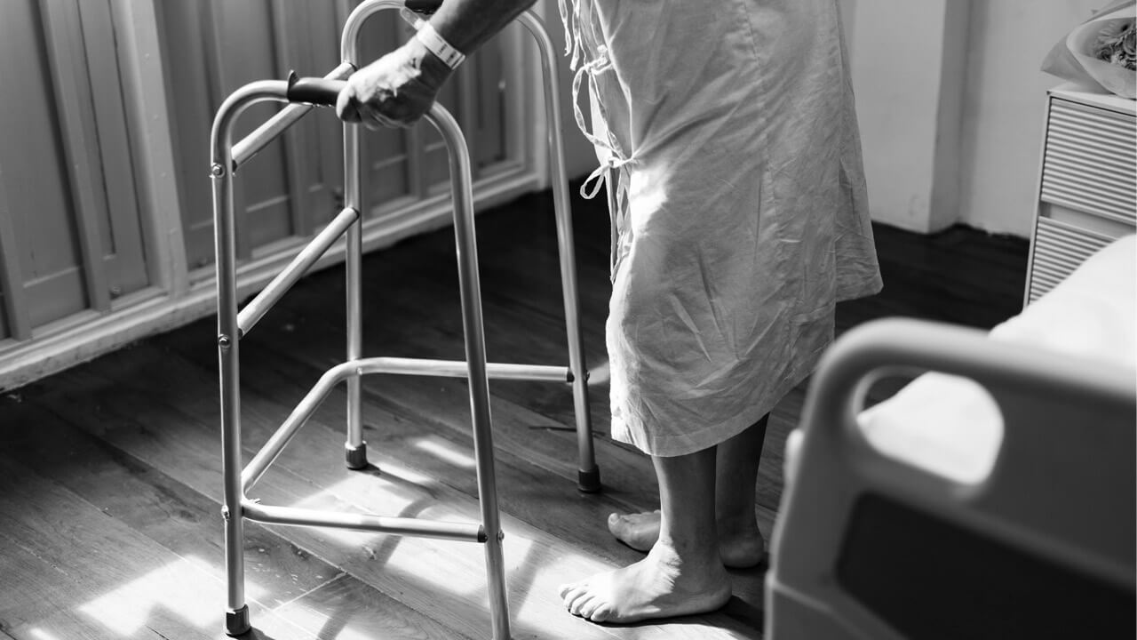 A person in a hospital gown, possibly experiencing early lupus symptoms, uses a walker for support while standing on a wooden floor. The person is barefoot and wearing a wristband, likely a hospital bracelet. A hospital bed is partially visible to the right side of the image.