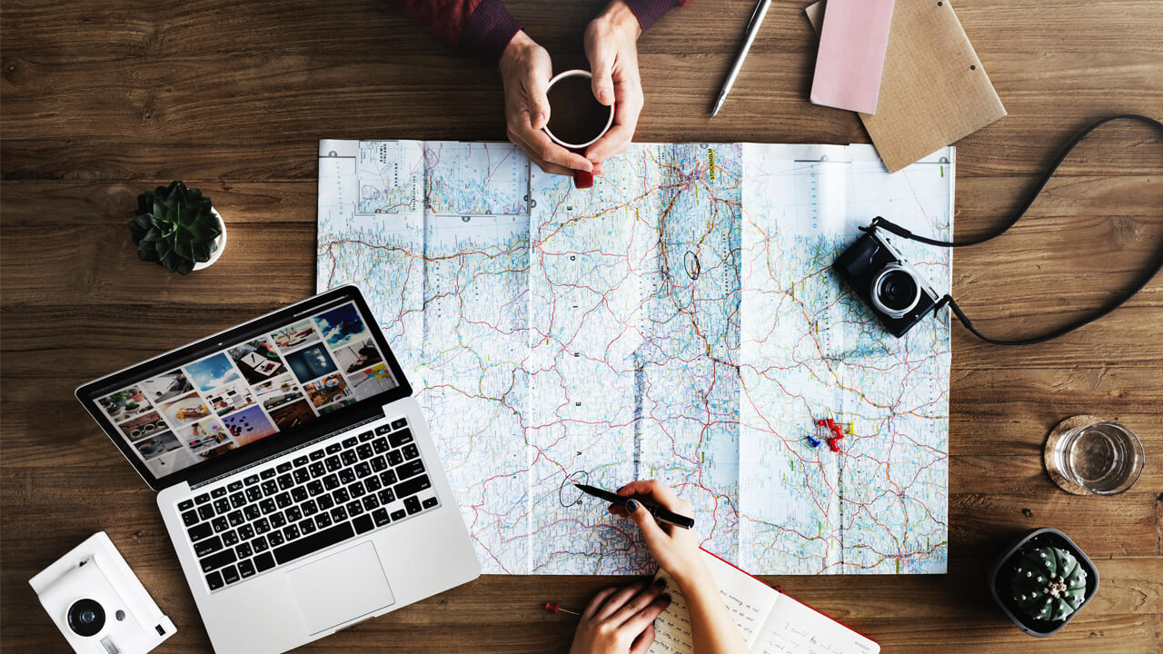 A wooden table with a large map covering most of it serves as a hub for holiday travel tips. Visible items include a laptop, camera, notepad, pen, instant camera, and a small plant. Two people are interacting with the map: one is holding a cup, and the other is writing on the map.