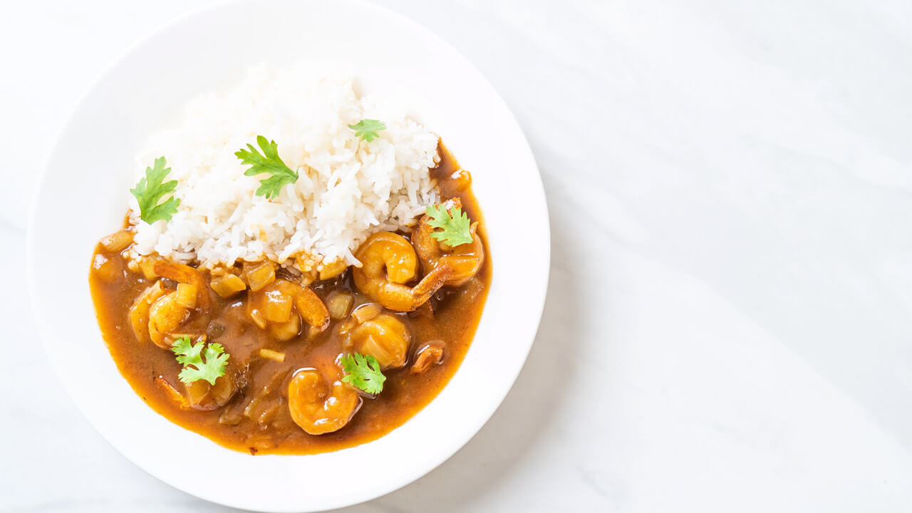 A white plate containing shrimp curry with a rich brown sauce and garnished with cilantro is served alongside a generous portion of steamed white rice. The dish is placed on a light-colored surface.