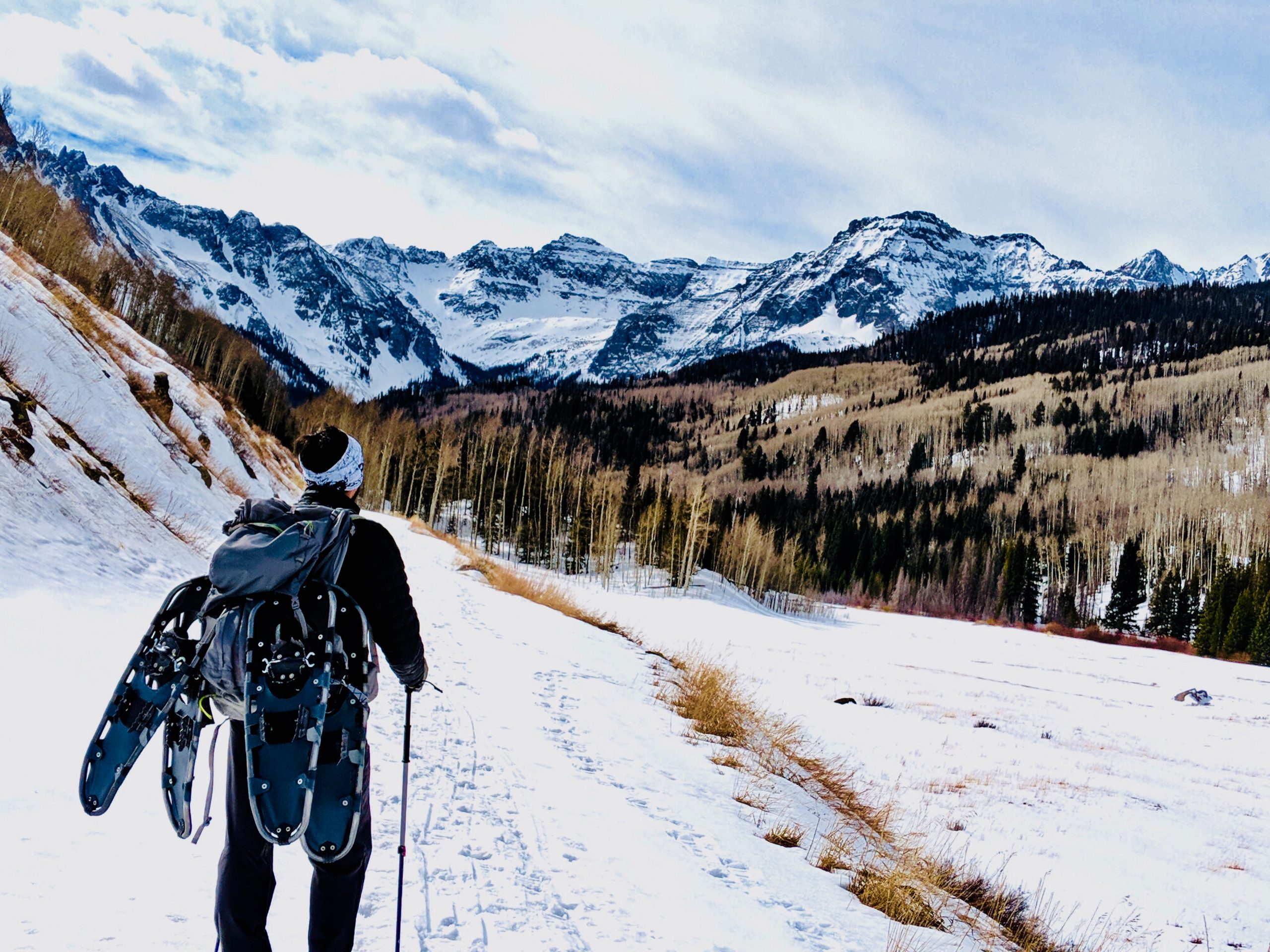A person carrying snowshoes on their backpack walks along a snowy path surrounded by leafless trees and snow-capped mountains under a cloudy sky.