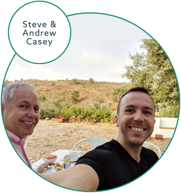 Two people smiling at the camera while sitting outdoors. One person on the left, older with grey hair, wears a pink shirt and holds a plate of food. The person on the right, younger with short hair, wears a black shirt. They are surrounded by trees and hills. Text reads "Steve & Andrew Casey.