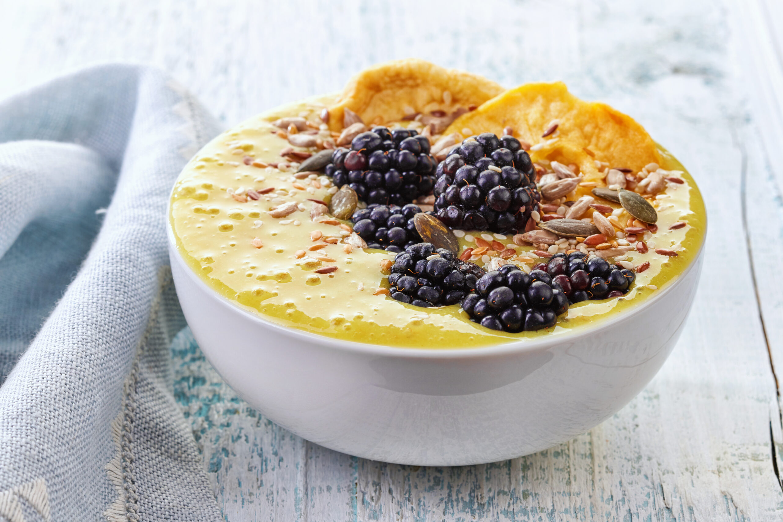 A bowl of creamy yellow smoothie topped with fresh blackberries, dried orange slices, sunflower seeds, and pumpkin seeds. The bowl is placed on a rustic wooden surface with a light blue cloth napkin to the side.