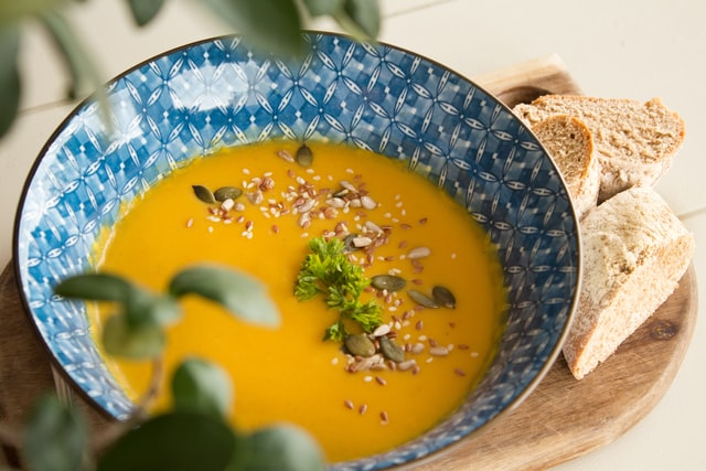 A blue patterned bowl filled with pumpkin soup garnished with seeds and a sprig of parsley. The bowl is placed on a wooden board with slices of bread on the side. A leafy plant is slightly visible in the foreground.
